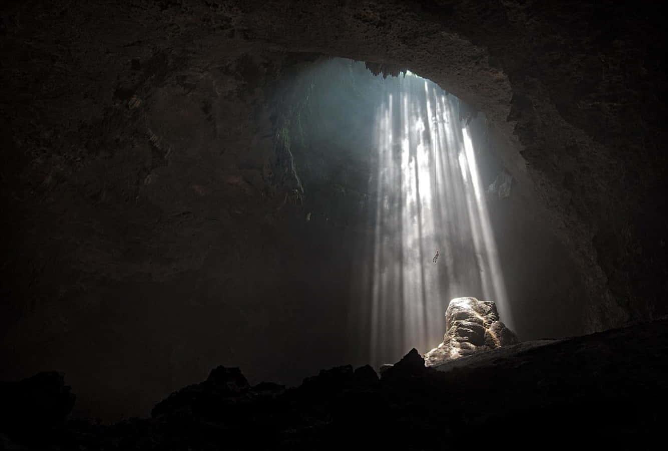 Cave 桌布 壁紙 — 陽光戲劇性地穿過洞穴開口，照亮下方孤獨的身影和崎嶇的岩石。