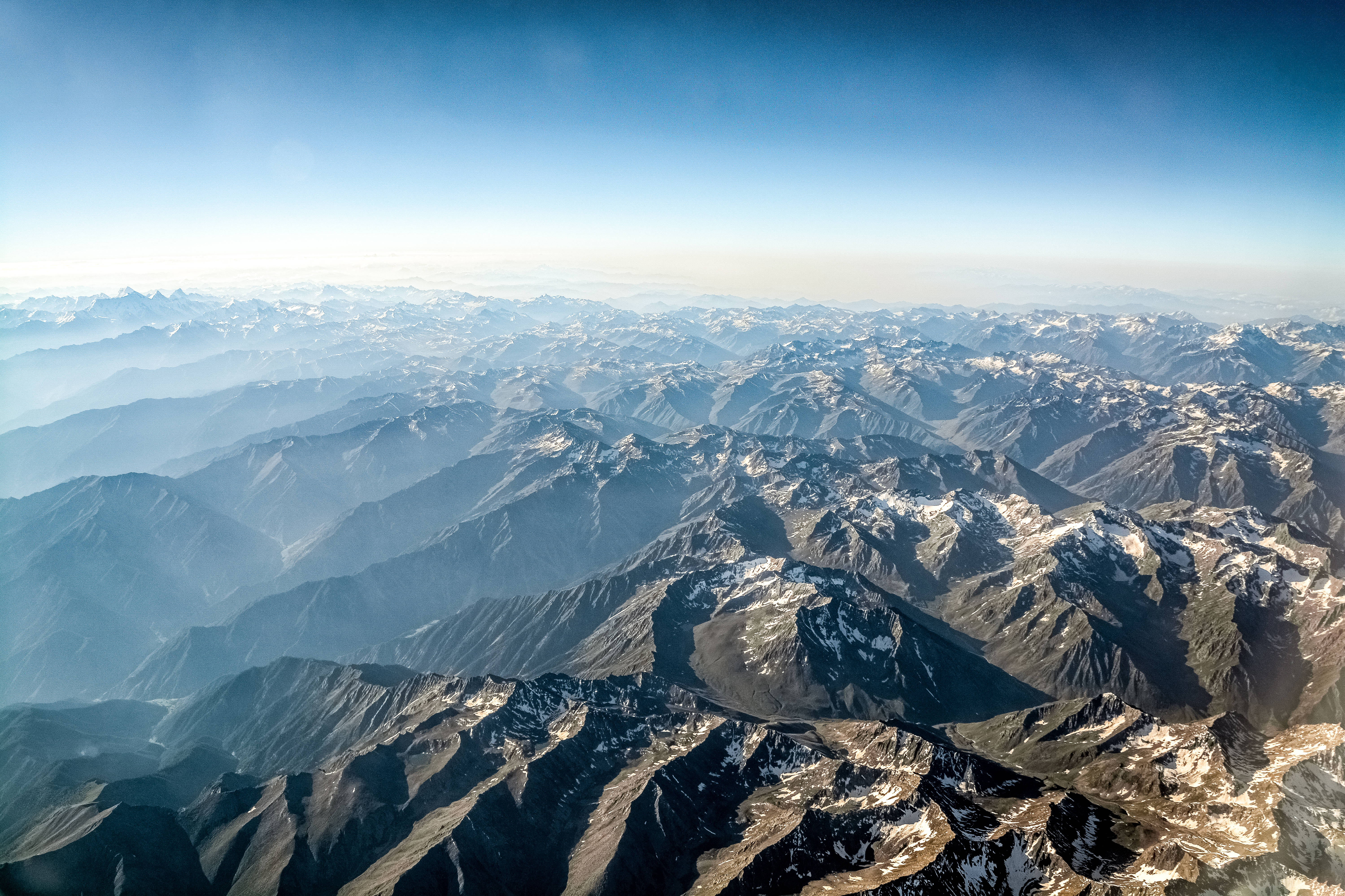 陽光揭開山地地形 壁紙 — 晴朗藍天下，無盡山脈的航拍視角，白雪覆蓋的山峰逐漸融入大氣薄霧中。