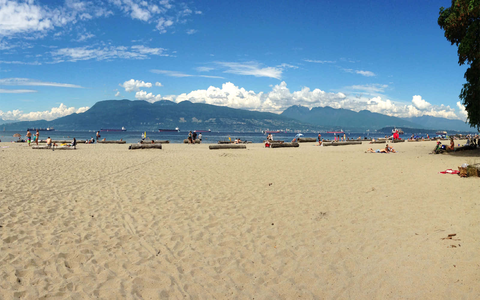Spanish Beach 桌布 壁紙 — 陽光明媚的海灘，人們在沙灘木頭上放鬆，天空湛藍，遠處有山脈和船隻。
