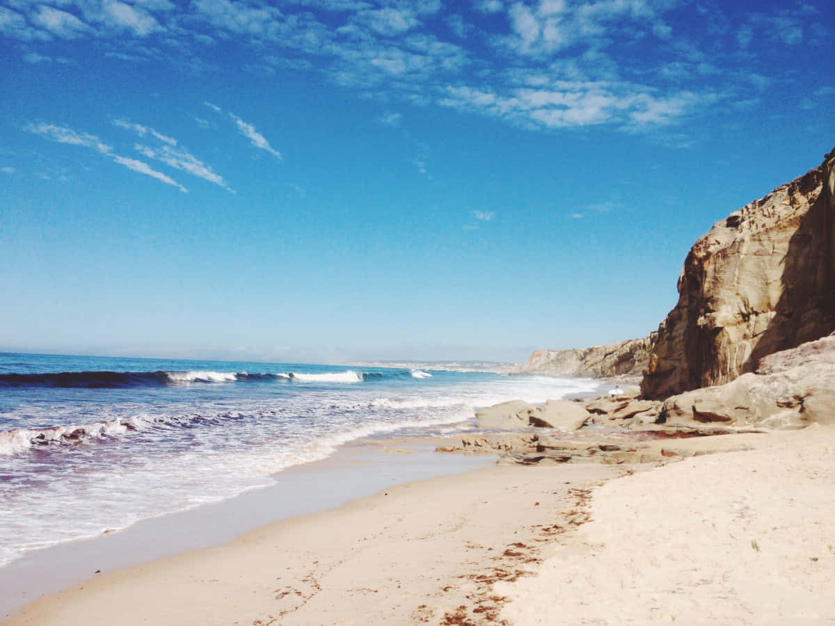 Beach Sand 桌布 壁紙 — 陽光明媚的海灘，在明亮的藍天下，碧綠色的海浪拍打著崎嶇懸崖旁的沙灘。