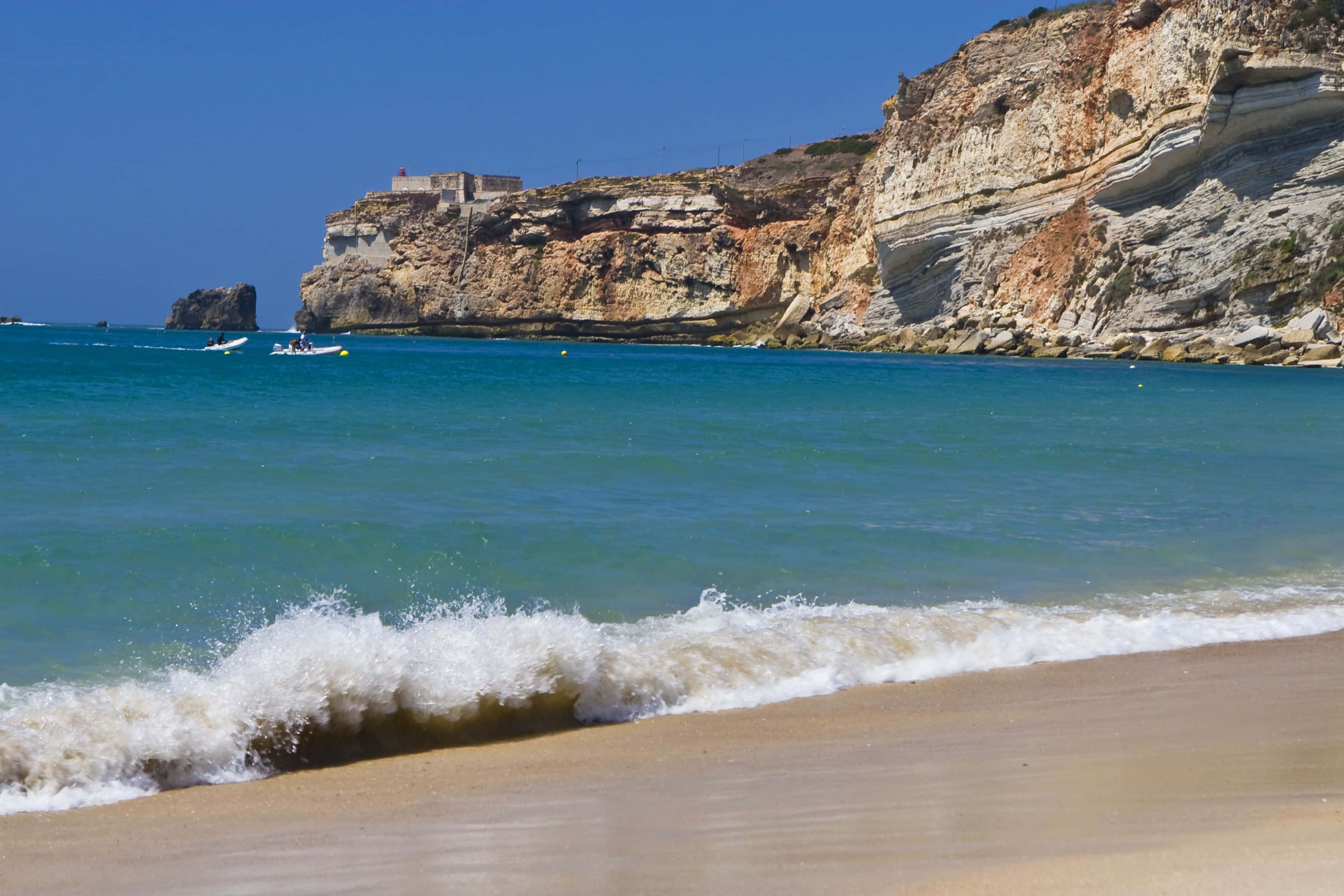 Portuguese Beach 桌布 壁紙 — 陽光明媚的海灘，在清澈的藍天下，層層疊疊的懸崖環繞著，綠松石色的海浪拍打著金色的沙灘。