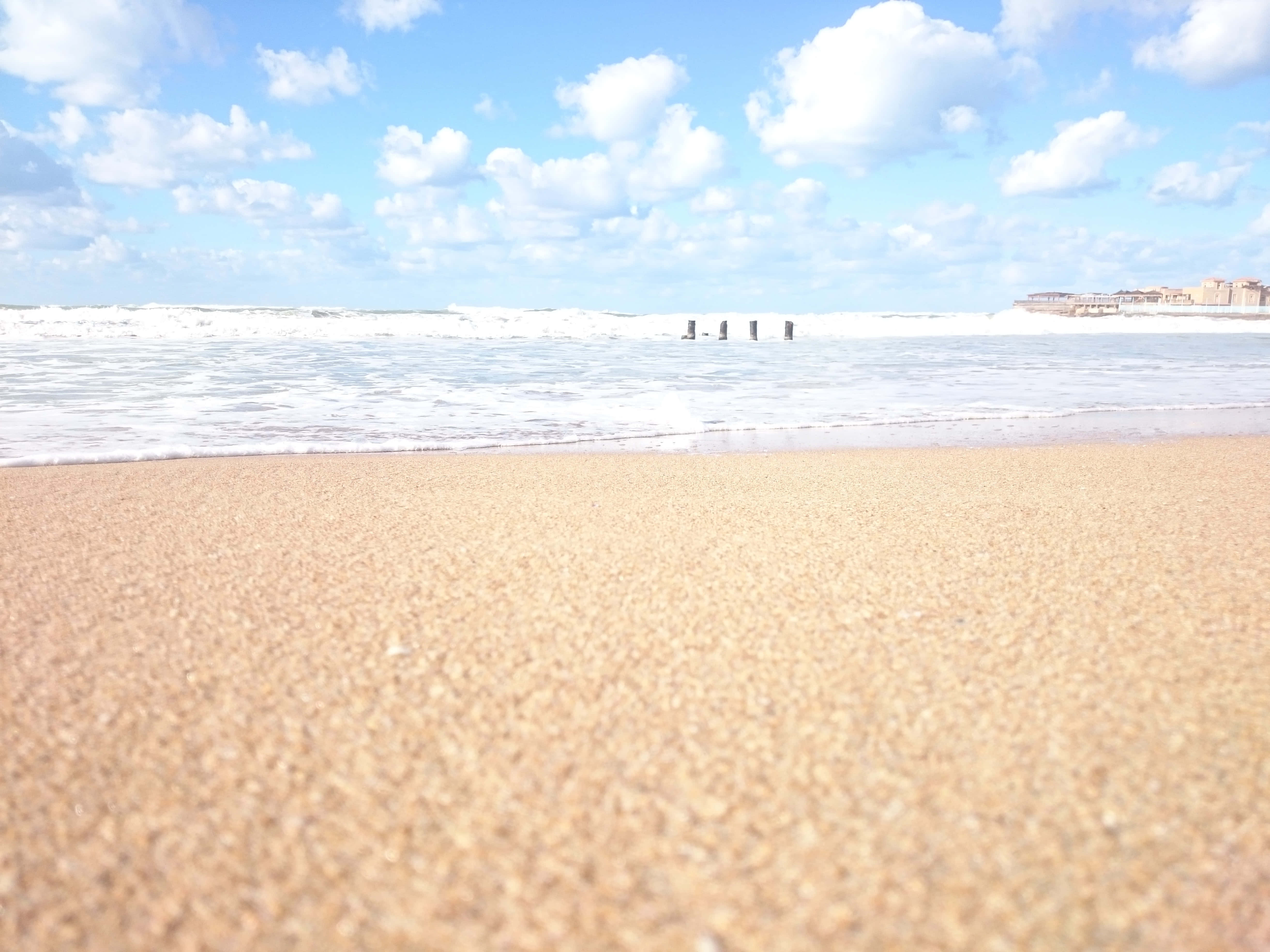 Beach Sand 桌布 壁紙 — 陽光明媚的海灘，金色沙灘與泡沫海浪交匯，湛藍天空點綴著蓬鬆白雲。