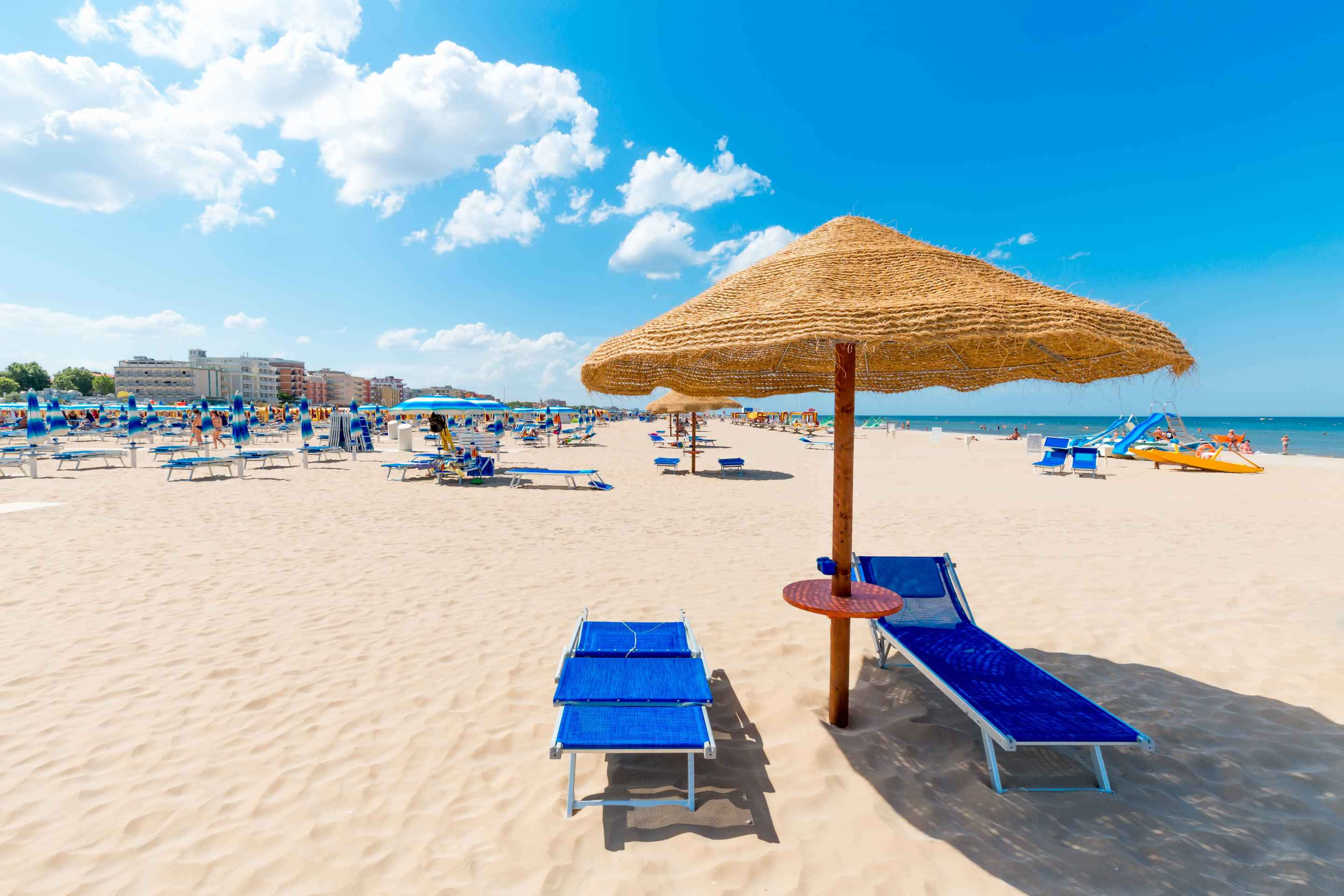 Italian Beach 桌布 壁紙 — 陽光明媚的海灘場景，藍色躺椅在草傘下，人們在明亮的藍天和蓬鬆的雲朵下享受海洋。