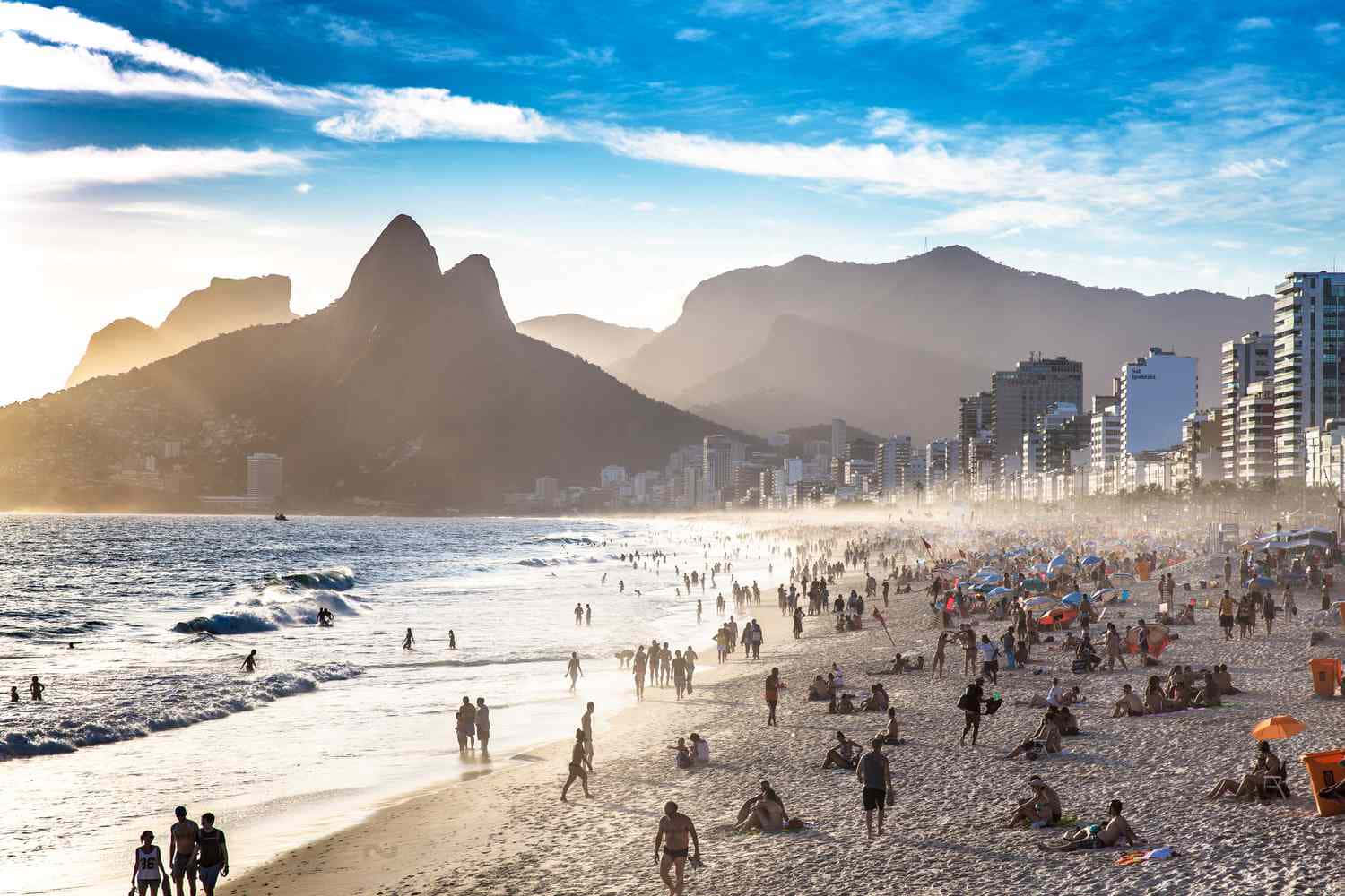 Brazilian Beach 桌布 壁紙 — 陽光明媚的里約熱內盧伊帕內瑪海灘上，人們在標誌性山脈和城市天際線的背景下享受海浪和沙灘，熱鬧非凡。