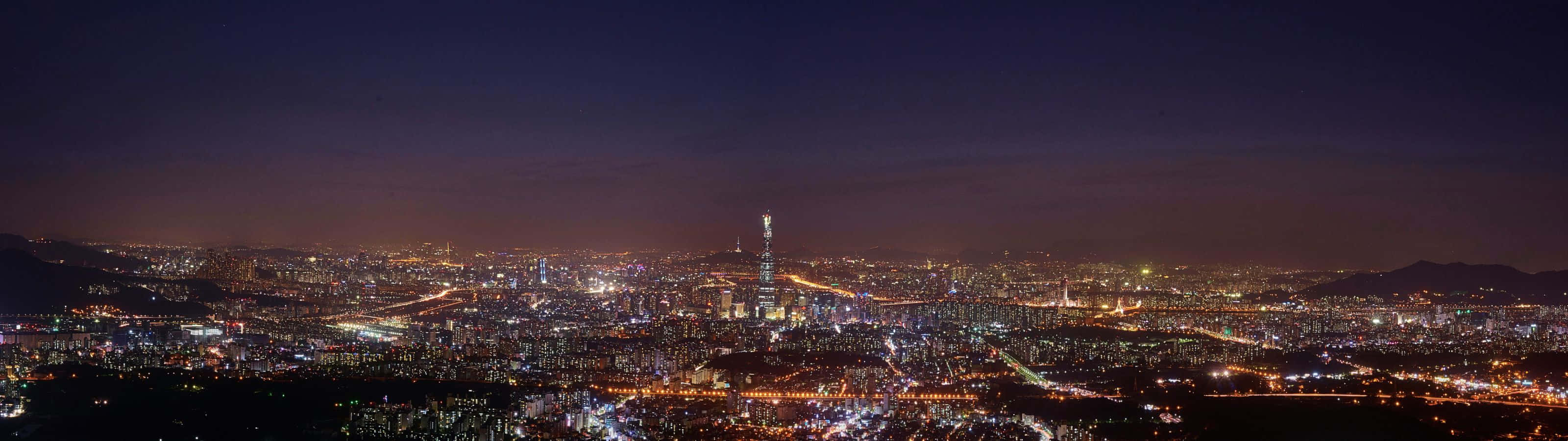 陽光普照下的純淨河谷與茂密森林 壁紙 — 廣闊城市景觀的 panoramatic 夜景，無數燈光點亮，高塔在黑暗天空中突出。