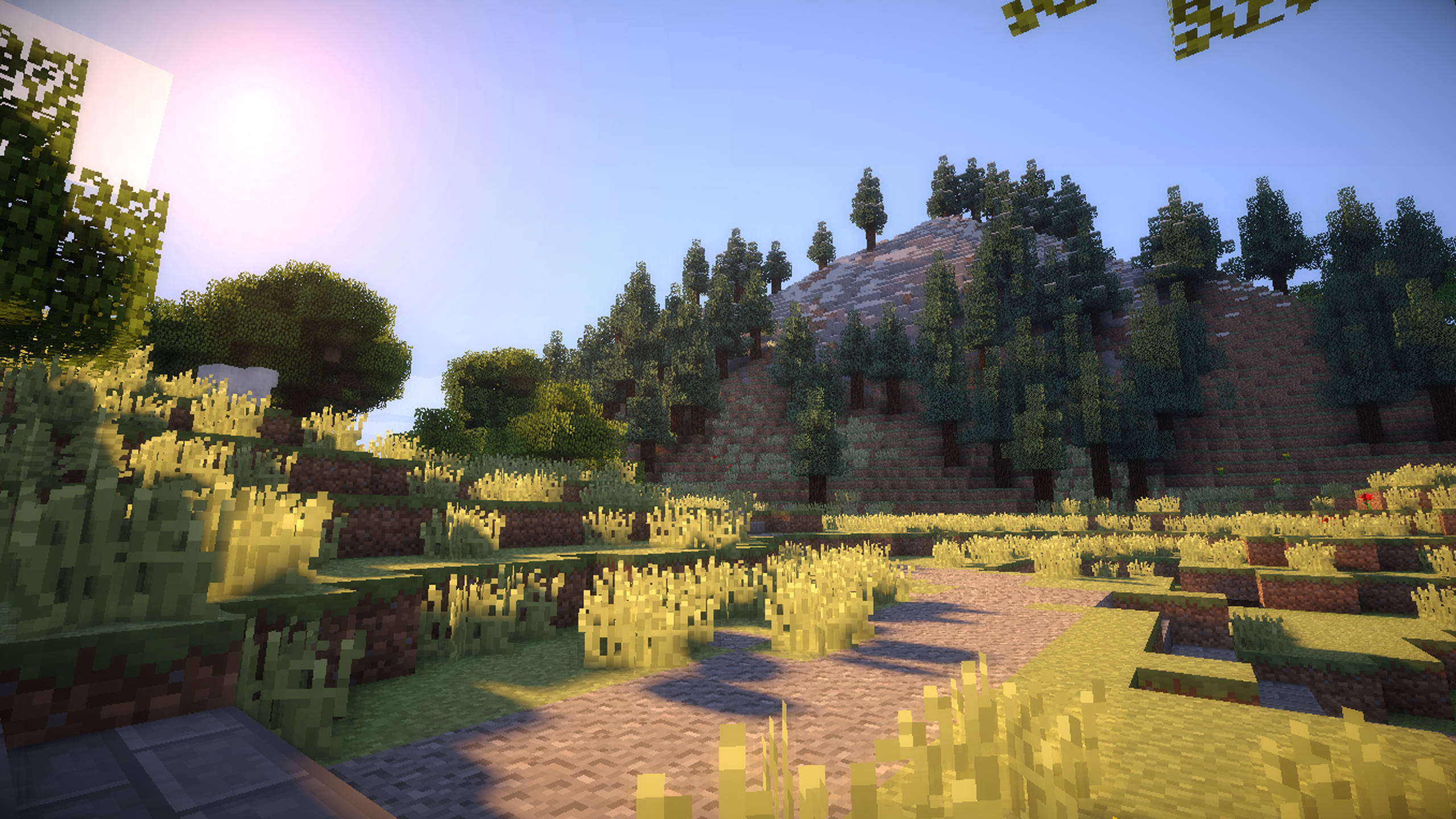 Minecraft Shaders 桌布 壁紙 — 陽光普照的像素化Minecraft景觀，有草地丘陵、樹木和一條石徑，在清澈的藍天下。