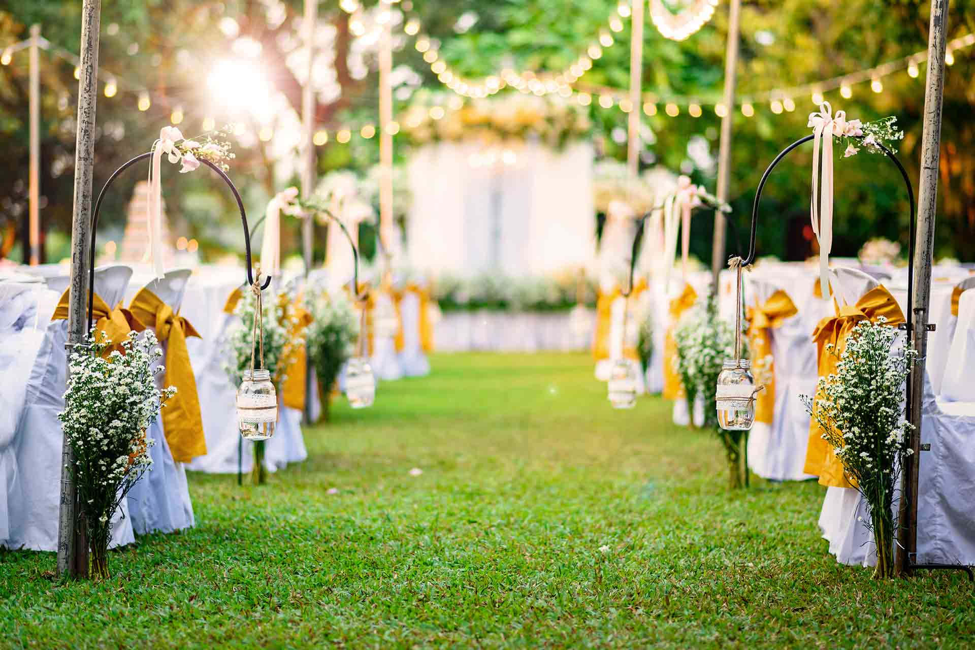 Garden Wedding 桌布 壁紙 — 陽光普照的戶外婚禮走道，兩旁擺放白色布藝椅子、金色蝴蝶結，以及掛在燈串下的燈籠。