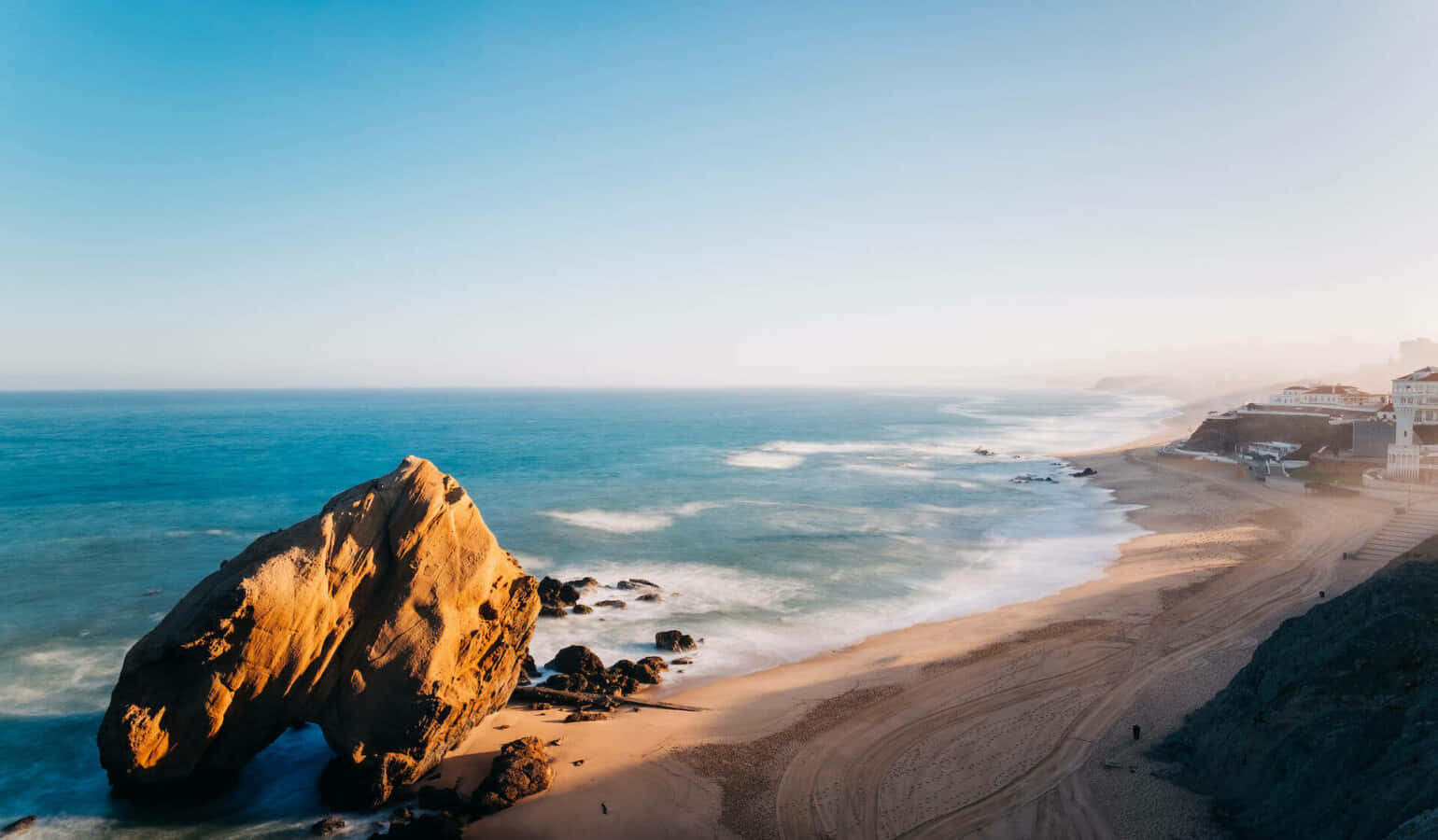 Portuguese Beach 桌布 壁紙 — 陽光普照的海岸景色，沙灘旁碧綠海水中有巨大的金色岩石突出。