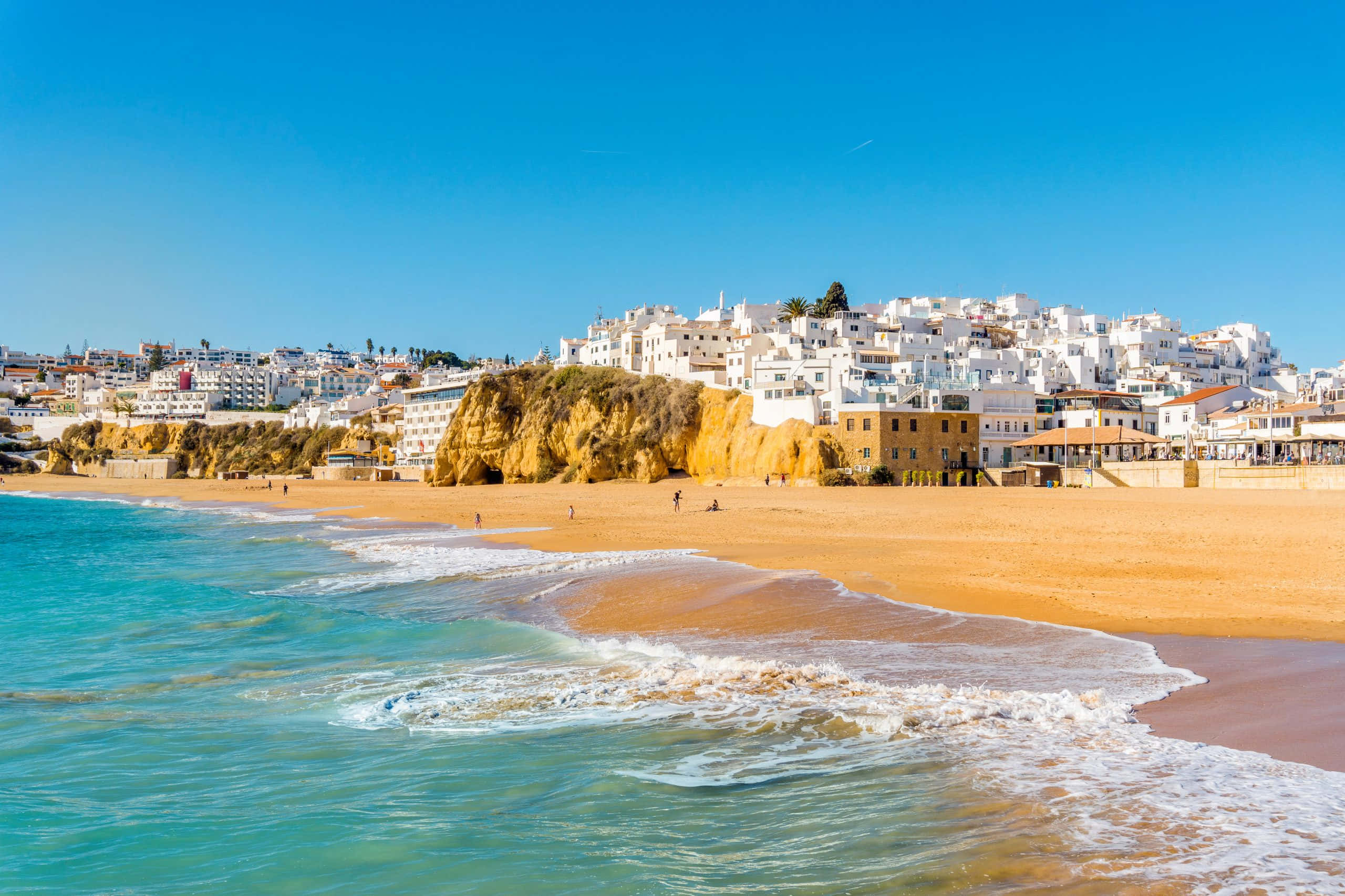 Portuguese Beach 桌布 壁紙 — 陽光普照的海灘，碧綠色的海浪拍打著金色沙灘，下方是白色懸崖小鎮，上方是清澈的藍天。