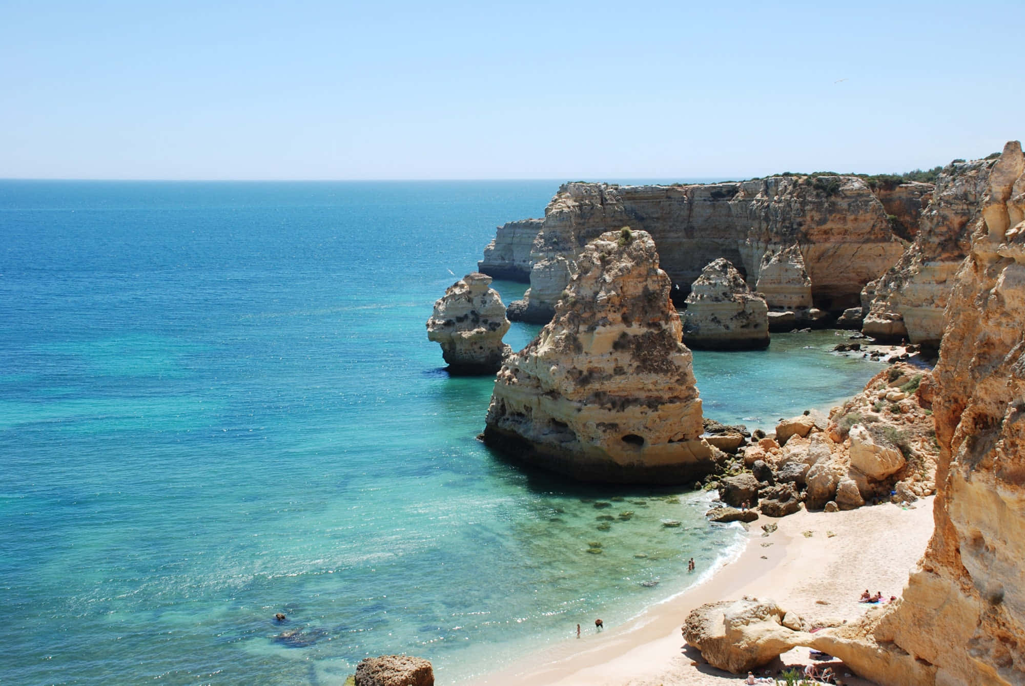 Portuguese Beach 桌布 壁紙 — 陽光普照的碧綠海水拍打著金色懸崖和沙灘，沙灘上有細小的人物享受海岸風光。