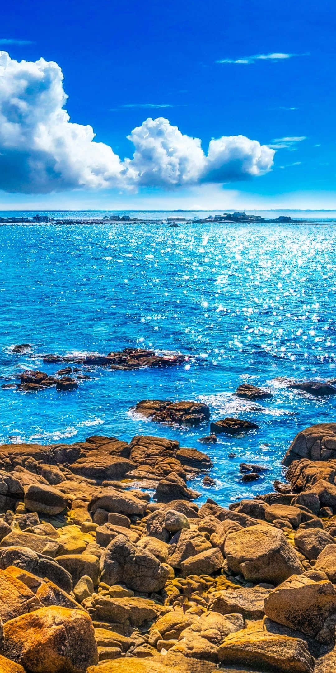 陽光普照的藍色海灘石景 壁紙 — 陽光在湛藍的海浪上舞動，拍打著金褐色的岩石海岸，天空湛藍，白雲朵朵。