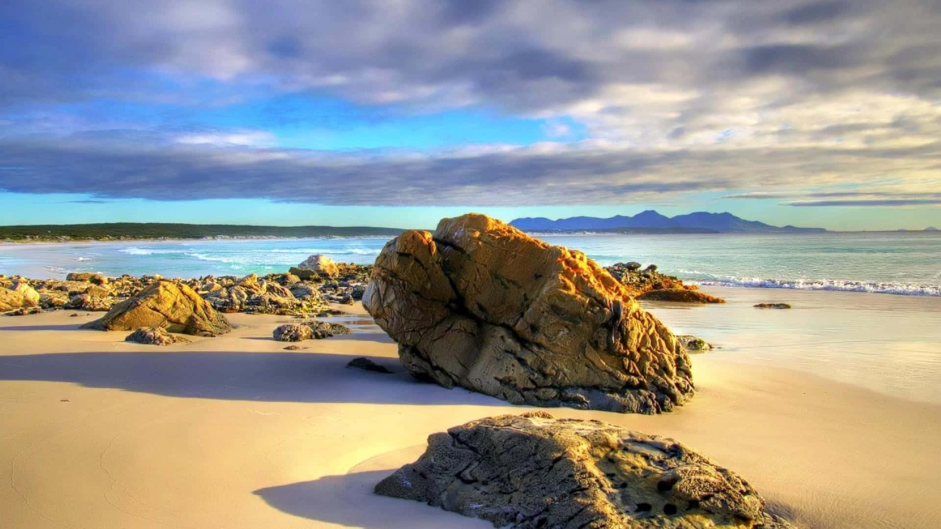 Australia Beach 桌布 壁紙 — 陽光普照的金色岩石點綴著寧靜的沙灘，溫柔的海浪在戲劇性的多雲天空下翻滾。