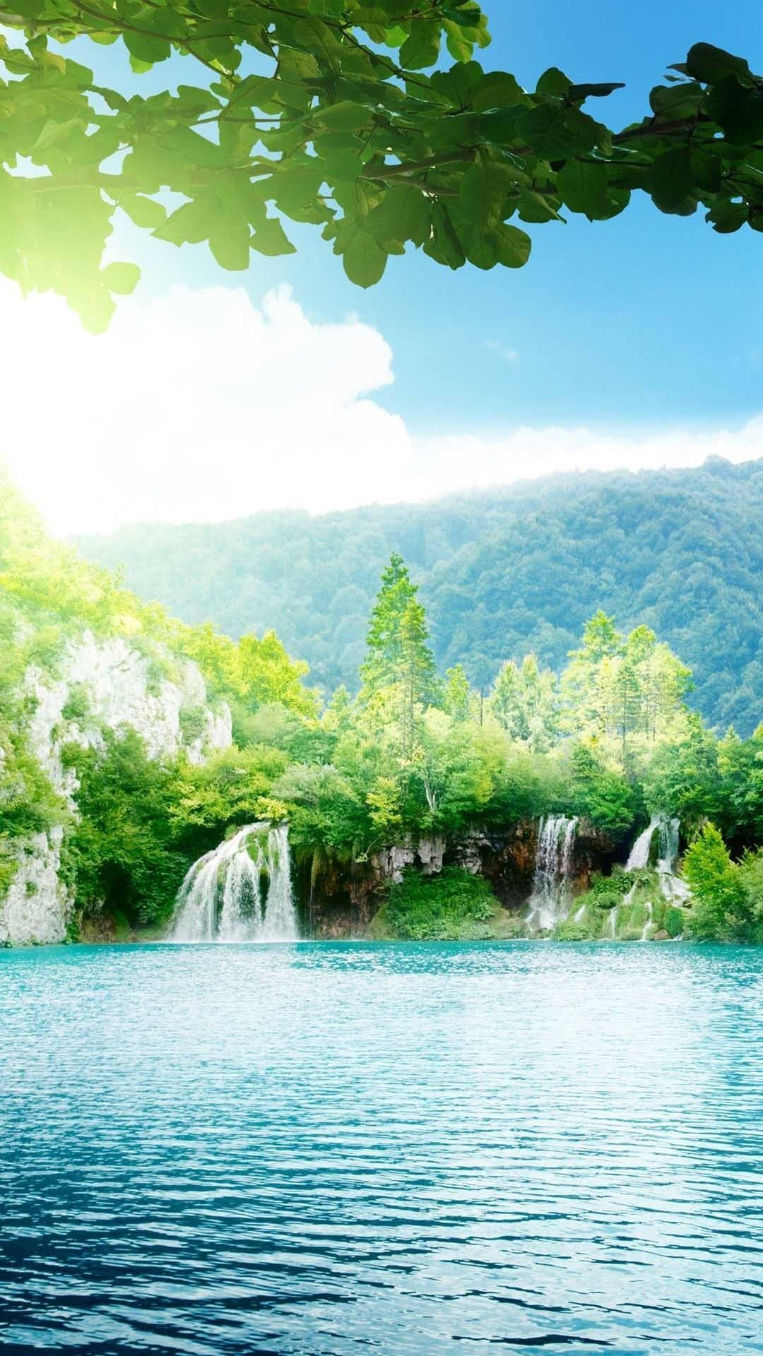 陽光瀑布風景 壁紙 — 陽光穿過茂密的綠葉，灑在寧靜的碧綠湖泊上，背景有層疊的瀑布和森林覆蓋的山丘。