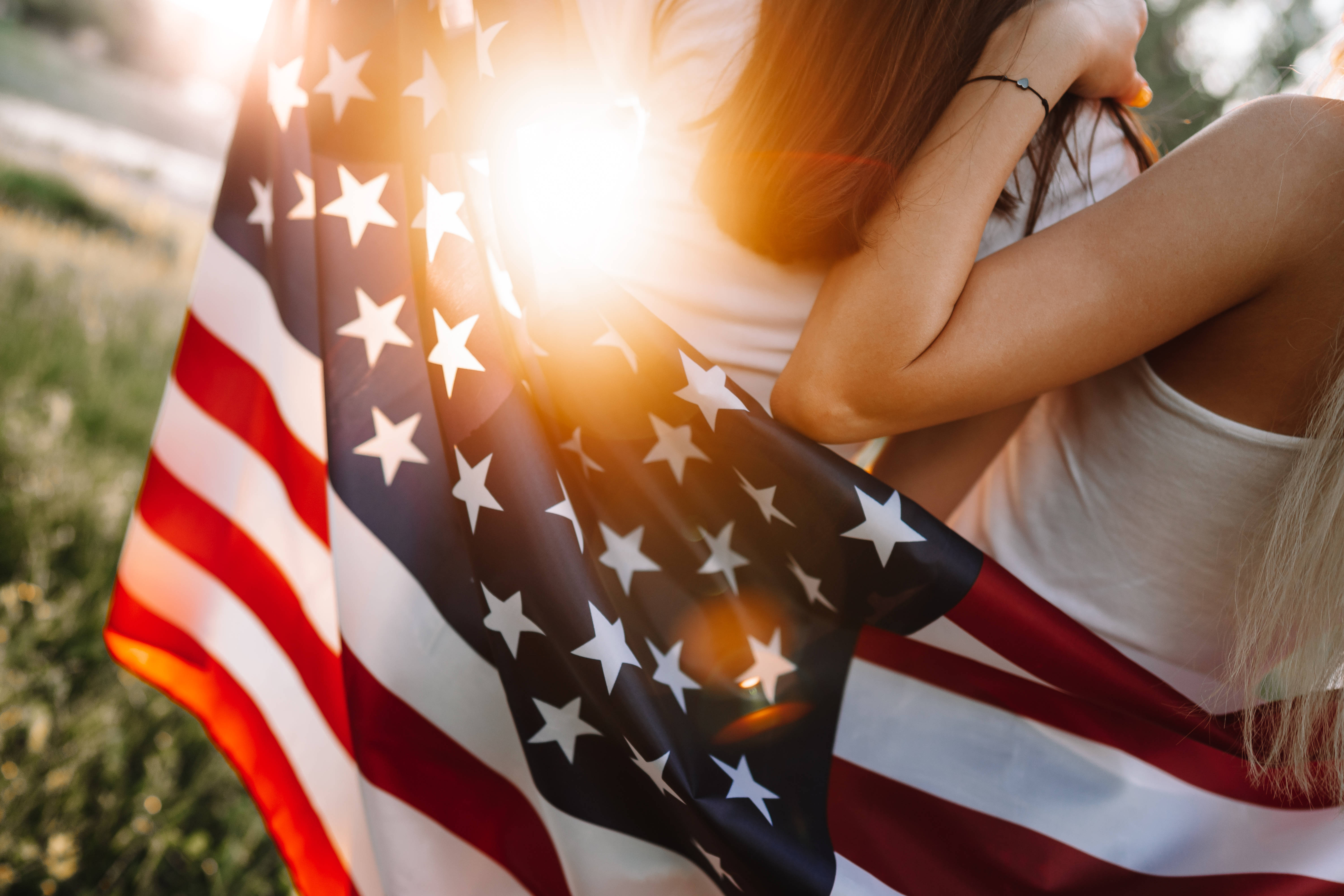陽光灑落美國國旗 iPhone 壁紙 — 一個人披著美國國旗擁抱自己，背後有太陽耀斑發光。