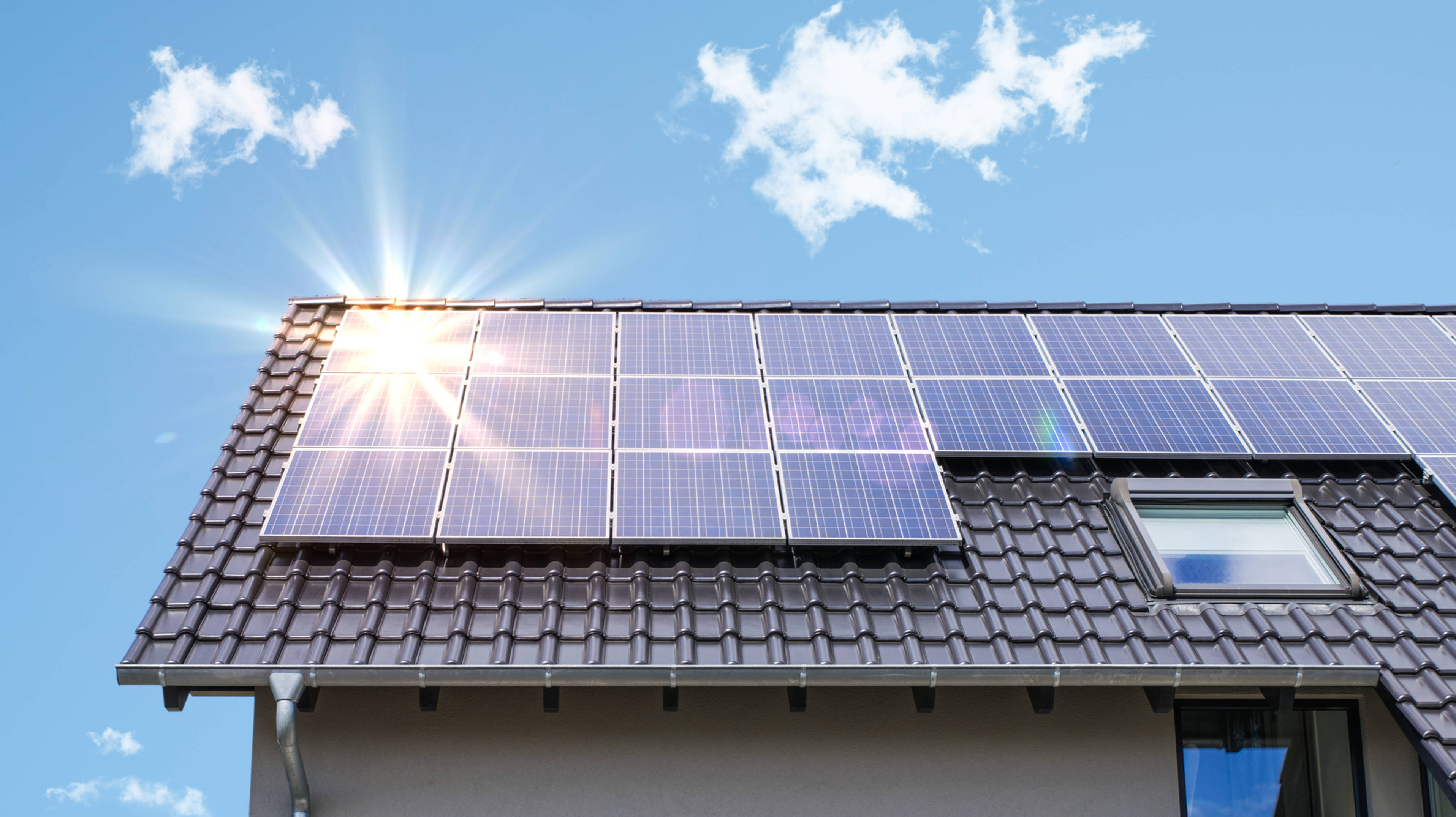 陽光照射NV Energy太陽能板 壁紙 — 太陽能電池板在明亮的陽光下在瓦片屋頂上閃閃發光，背景是清澈的藍天和縷縷白雲。