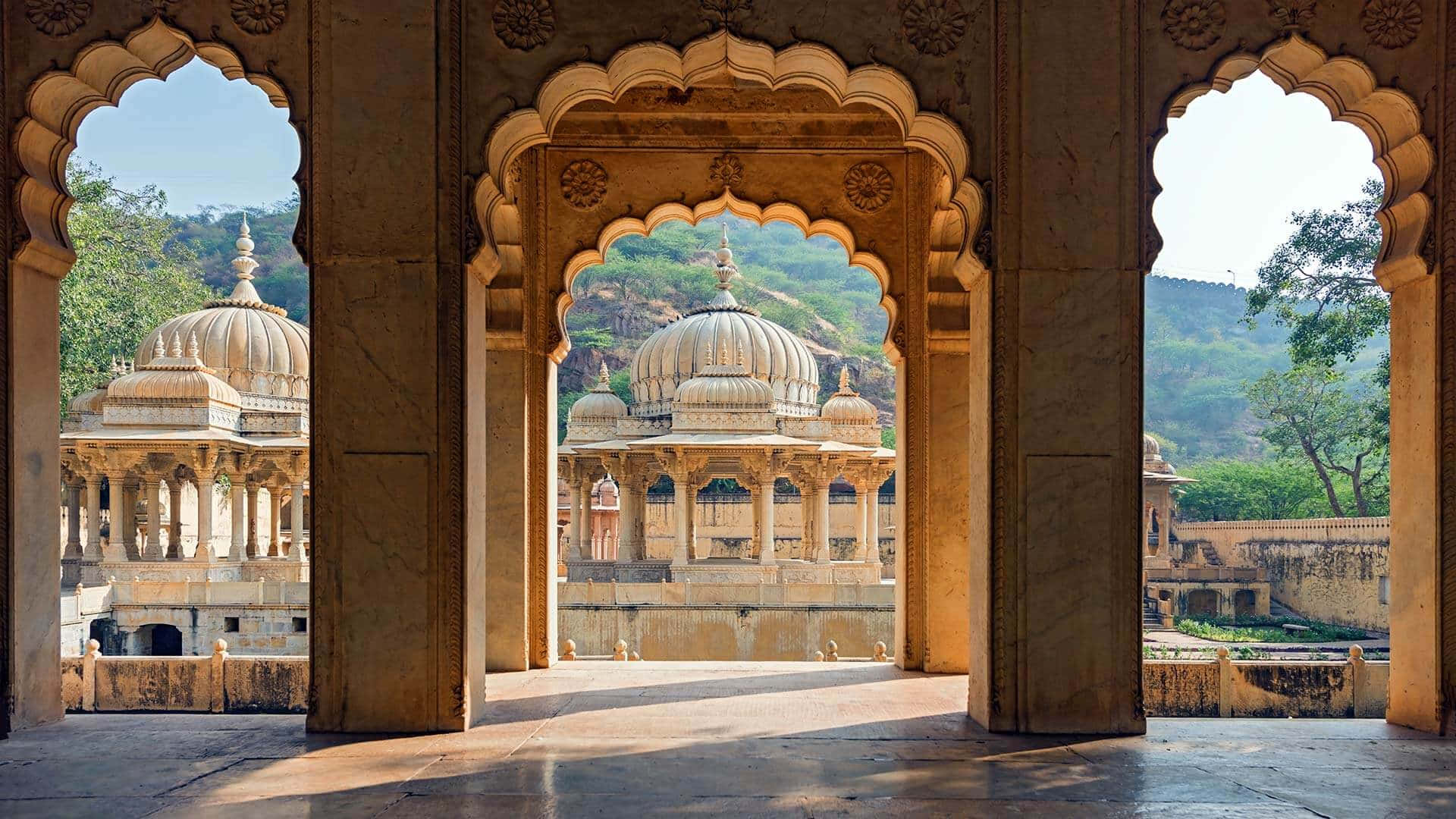 Rajasthan 桌布 壁紙 — 陽光照射的石拱門框住一座雄偉的圓頂宮殿，背景是鬱鬱蔥蔥的綠色山丘，天空清澈湛藍。