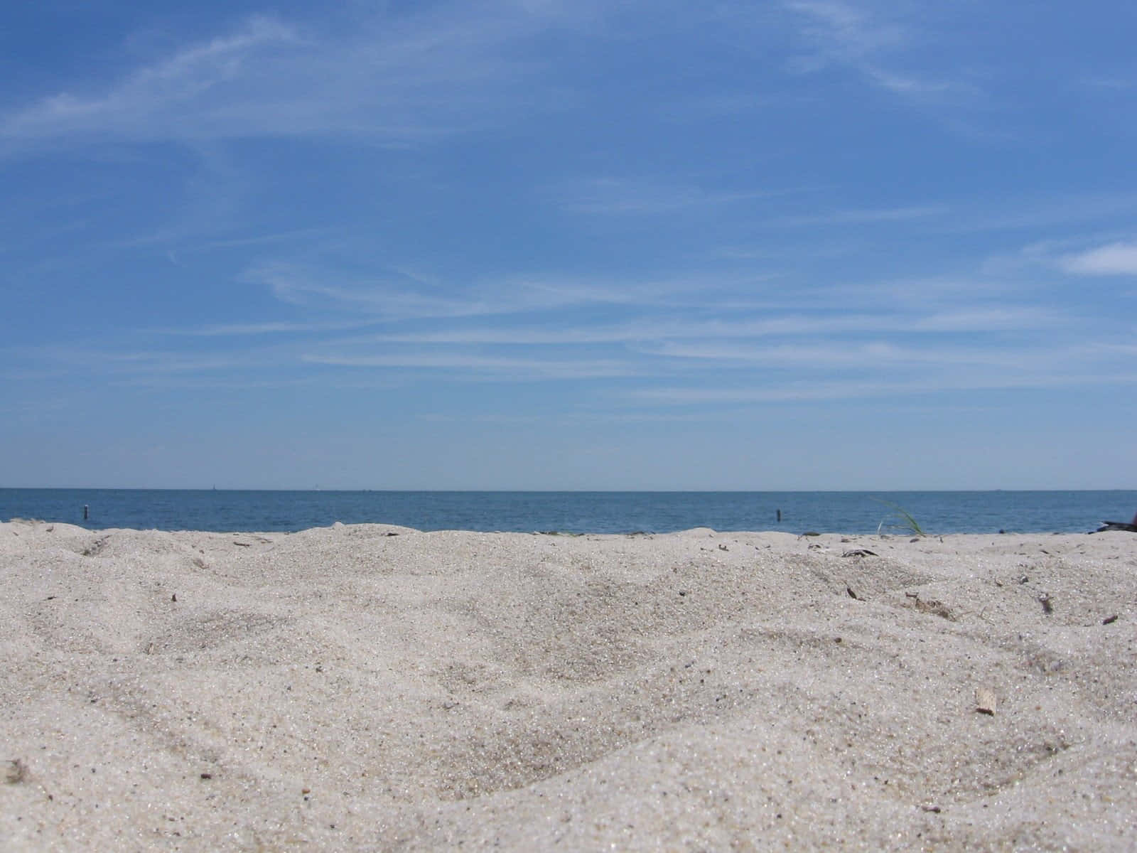 Beach Sand 桌布 壁紙 — 陽光照耀的沙灘在晴朗的天空下延伸至平靜的藍色海洋，天空中有縷縷雲彩。