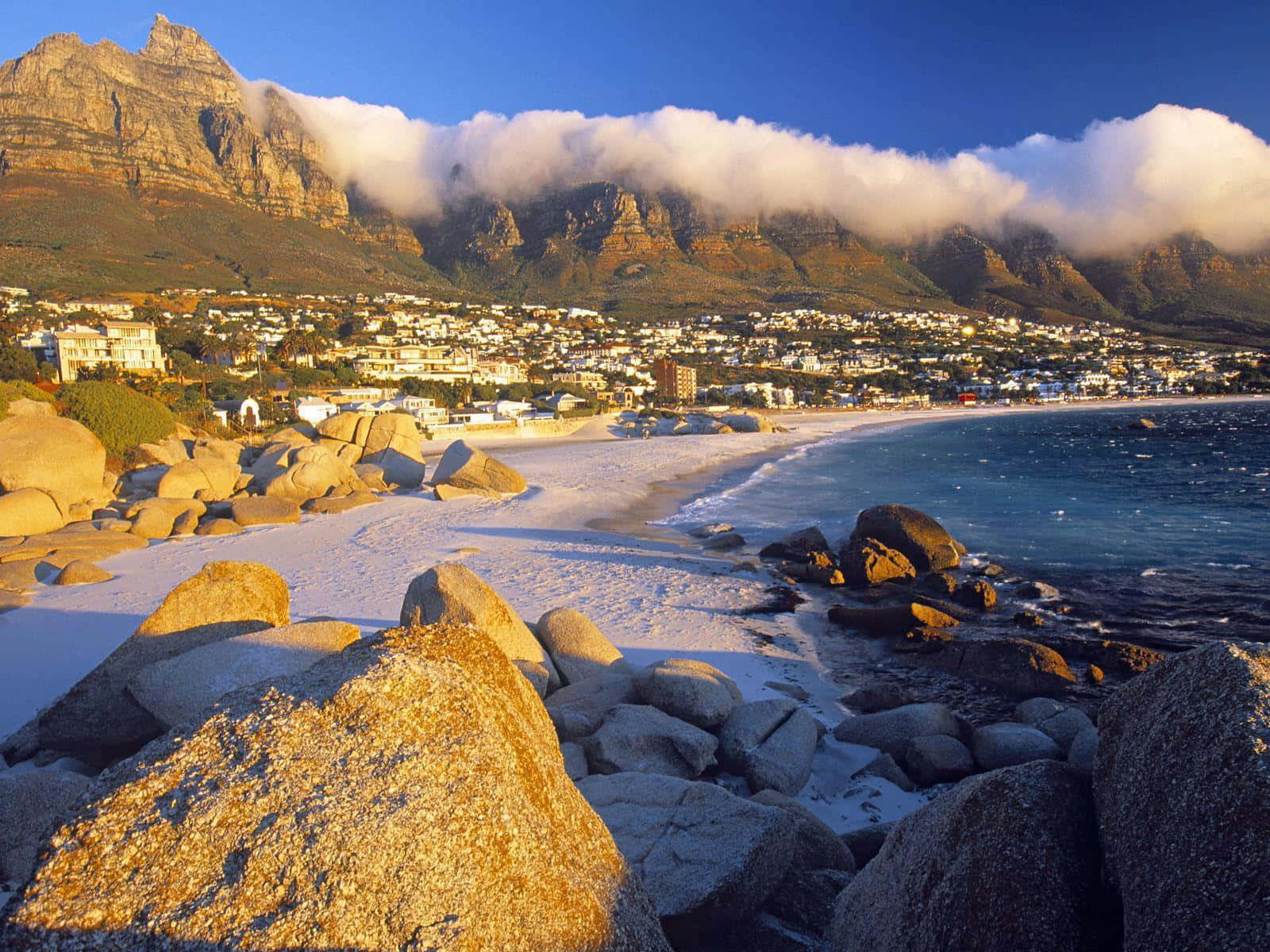 South Africa 桌布 壁紙 — 陽光照耀的海灘，前景有巨大圓石，海岸小鎮後方有被雲層部分遮蔽的壯麗山脈。