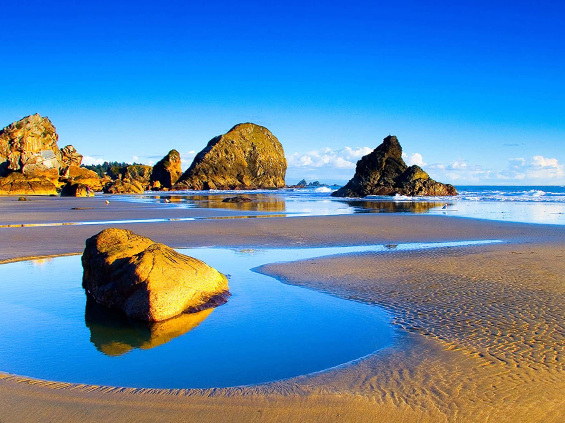 Beautiful Beach 桌布 壁紙 — 陽光照耀的海灘，金色沙紋和巨石在清澈藍天下倒映在潮汐池中。