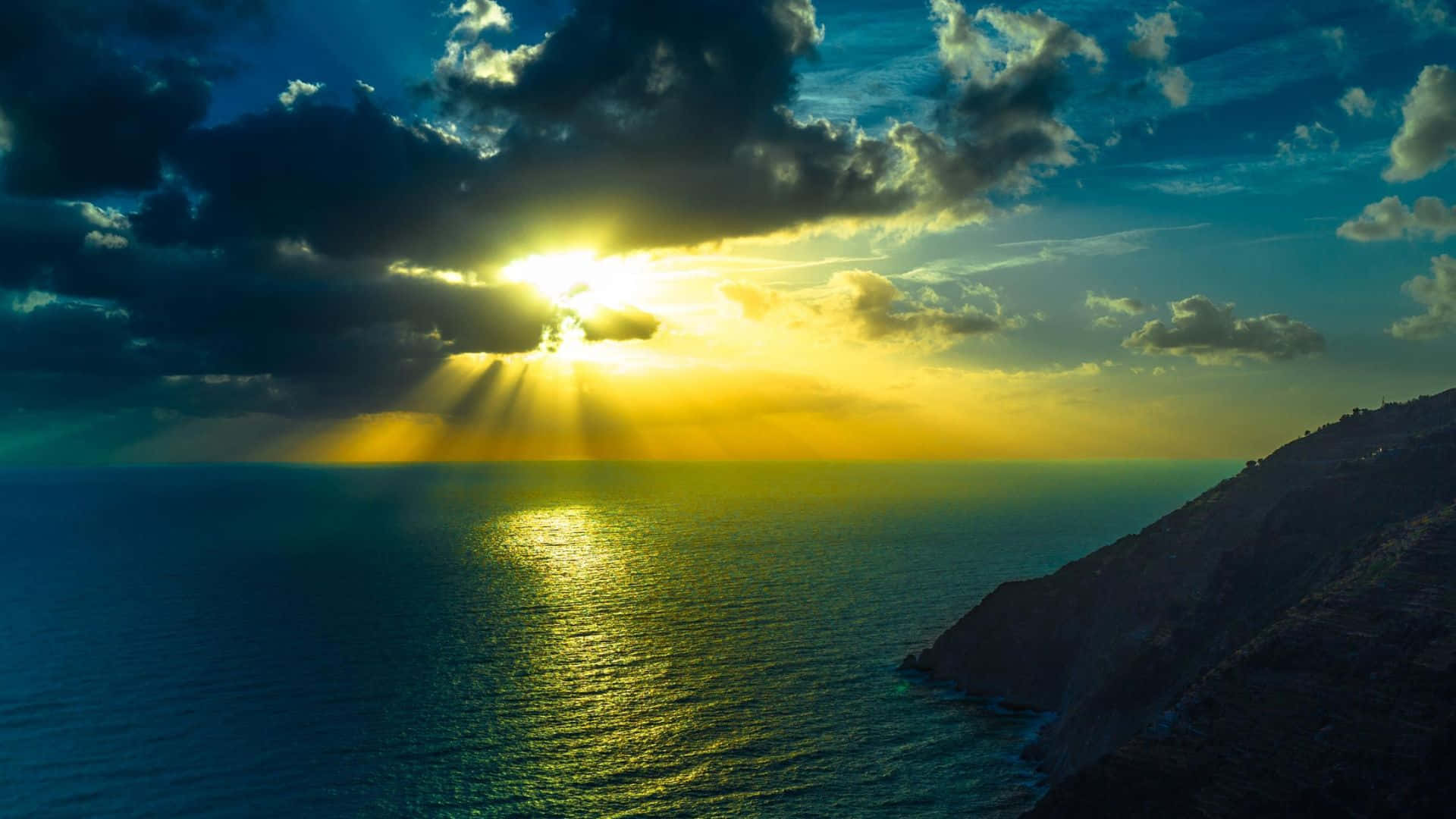 Beautiful Ocean 桌布 壁紙 — 陽光穿透廣闊海洋上空的烏雲，在崎嶇懸崖旁的深藍色海面上投下金色光芒。
