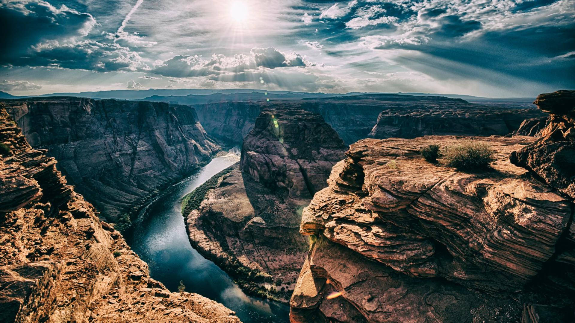 Arizona 桌布 壁紙 — 陽光穿透戲劇性的雲層，照耀在一條蜿蜒穿過深紅色岩石峽谷的河流上。