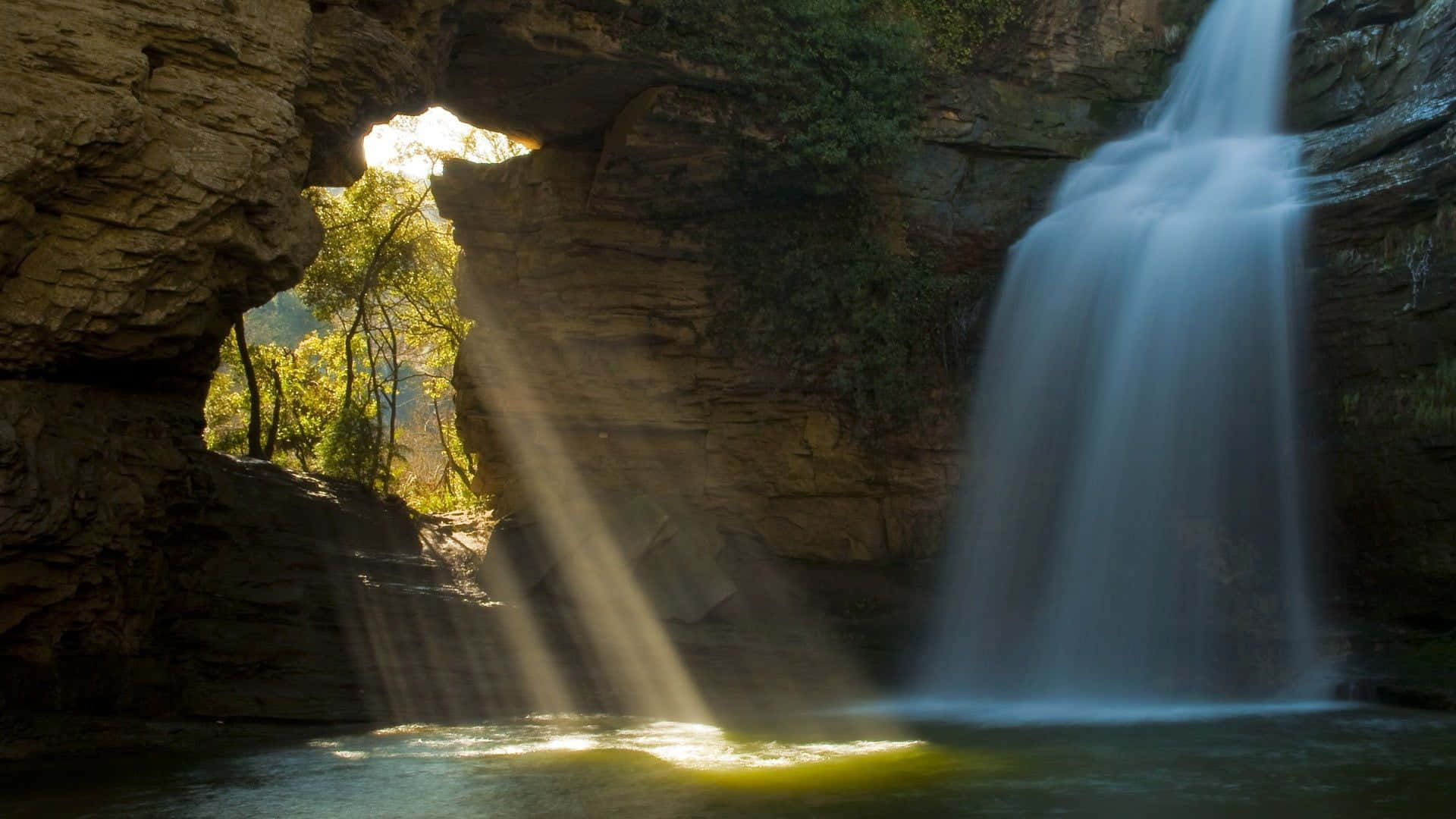 Waterfalls 桌布 壁紙 — 陽光穿過天然岩石拱門，照亮了層疊的瀑布和長滿苔蘚的懸崖。