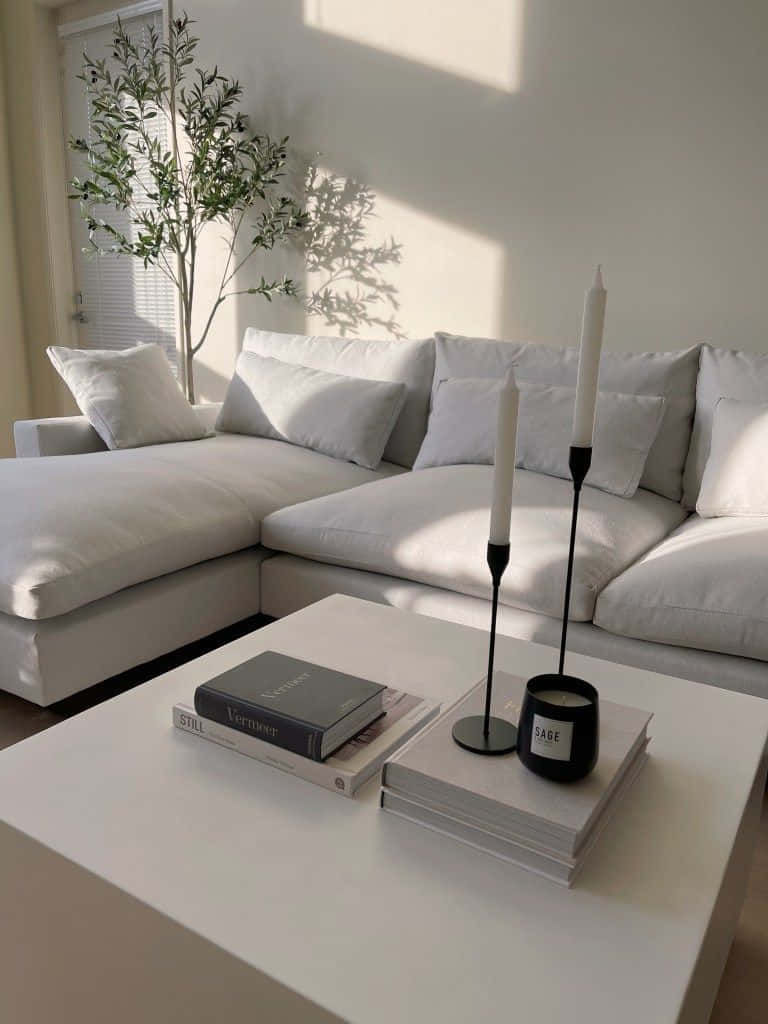 Minimalist Design 桌布 壁紙 — 陽光穿過百葉窗，在白色組合沙發上投下葉影，旁邊有一棵高大的盆栽植物和簡約的咖啡桌，桌上放著書籍、蠟燭和鼠尾草蠟燭。
