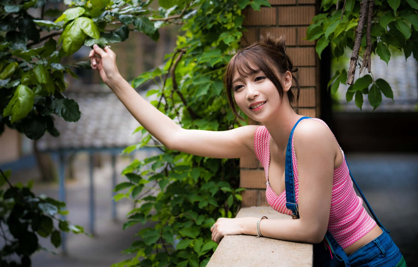 陽台上的可愛亞洲女孩 壁紙 — 一位微笑的年輕女子身穿粉紅色上衣和牛仔工人褲，靠在石台上，輕輕觸摸磚牆旁茂密的綠葉。