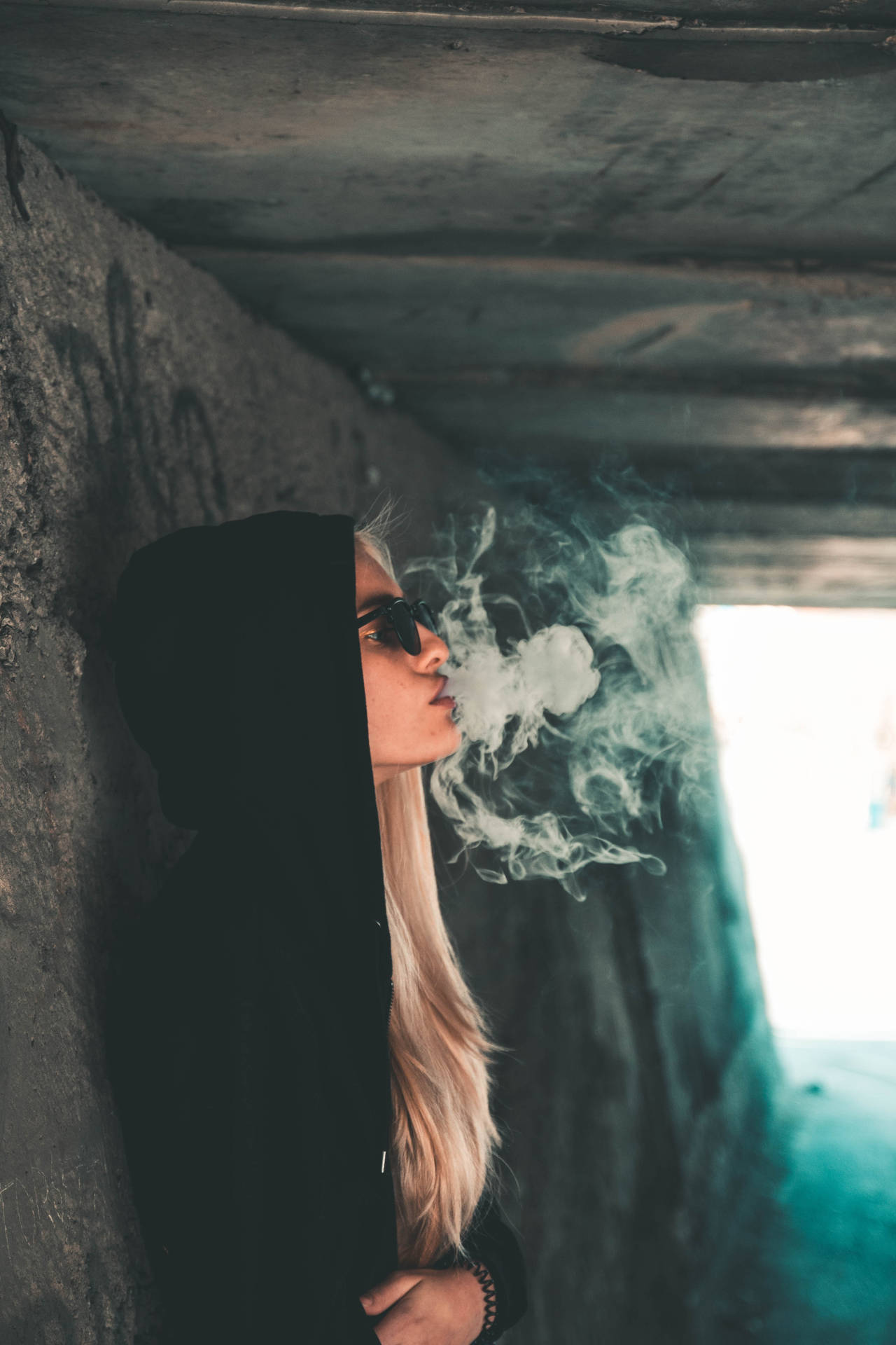 穿著黑色連帽衫和太陽眼鏡的人，在一個佈滿塗鴉的地下通道中呼出煙霧。