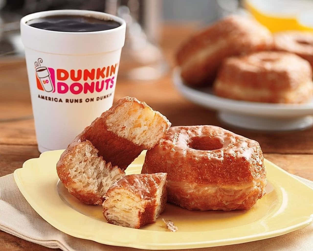 隨時享用Dunkin' Donuts美味甜甜圈！ 壁紙 — 一個溫暖的糖霜冬甩被掰開一半，放在一個黃色碟上，旁邊有一杯Dunkin' Donuts咖啡。