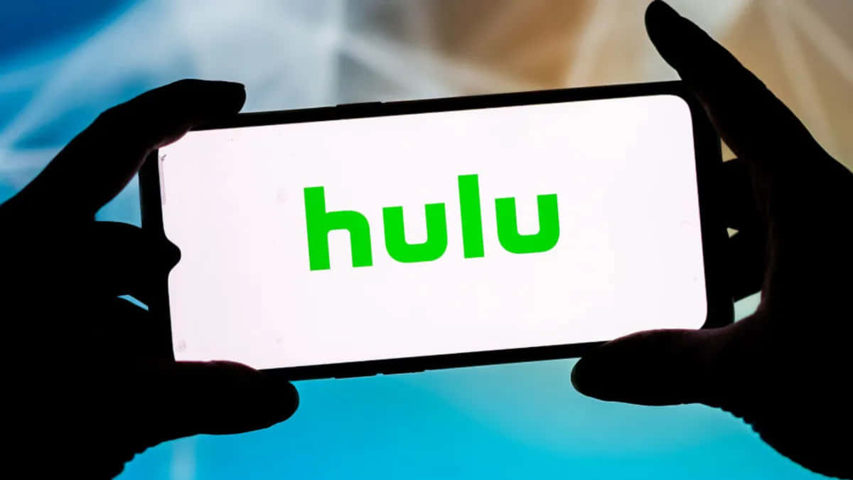 隨時掌握 Hulu 最新動態 壁紙 — 剪影的手拿著一部智能手機，在模糊的藍色背景上顯示鮮綠色的 Hulu 標誌。
