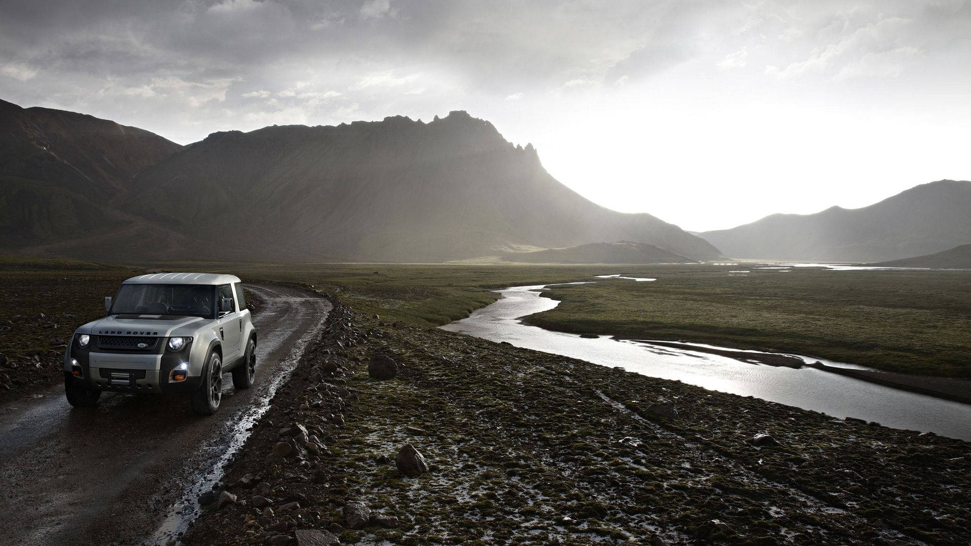 銀色 Land Rover Defender 在日落時分靠近河流的泥濘小路上，背景有山脈。
