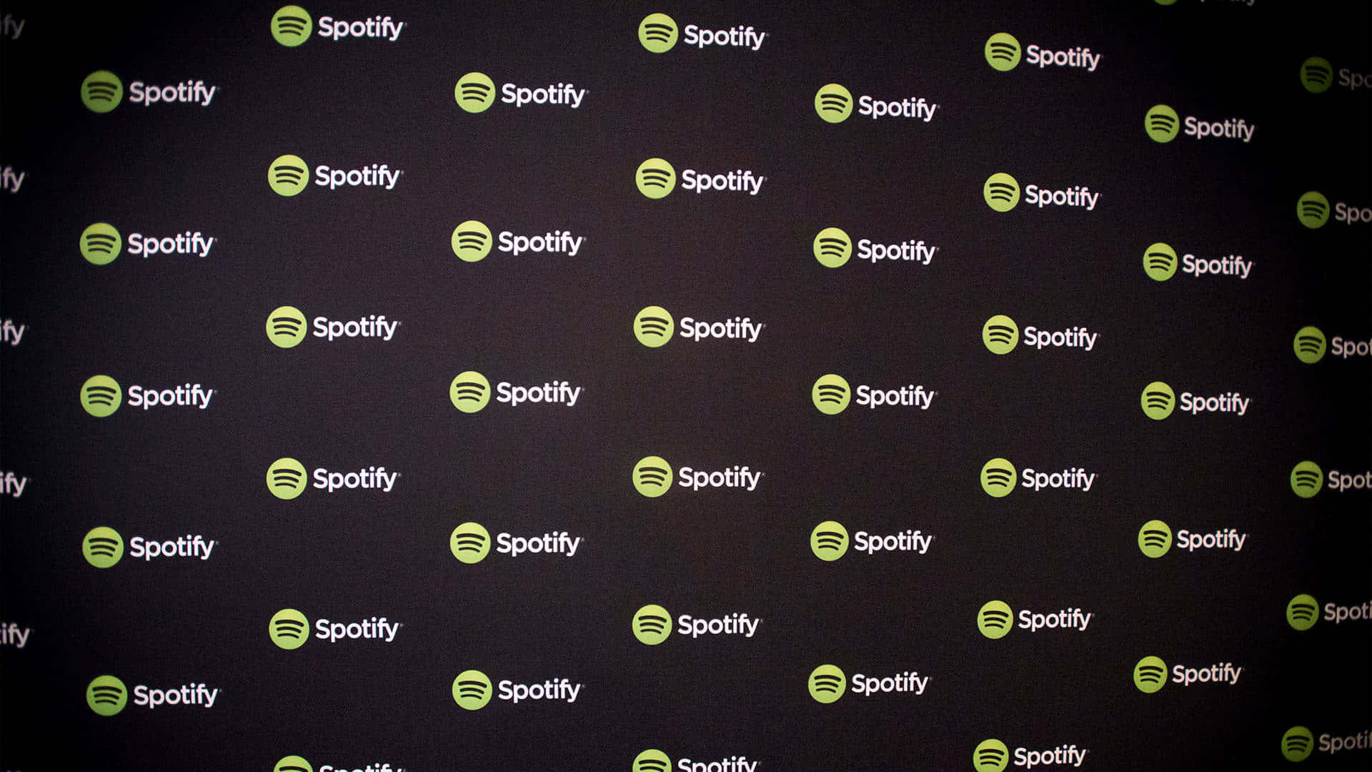 隨時隨地享受最新音樂與播客，盡在Spotify 壁紙 — 一面深色牆壁覆蓋著重複的綠色和白色Spotify標誌，形成品牌圖案。