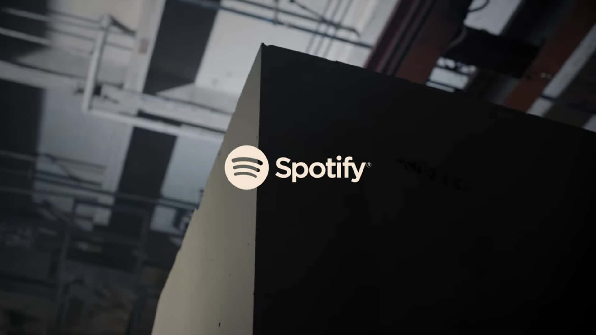 隨時隨地聆聽你最愛的藝人 壁紙 — 特寫鏡頭：Spotify 標誌在黑色表面上發出白色光芒，背景是模糊的工業環境。