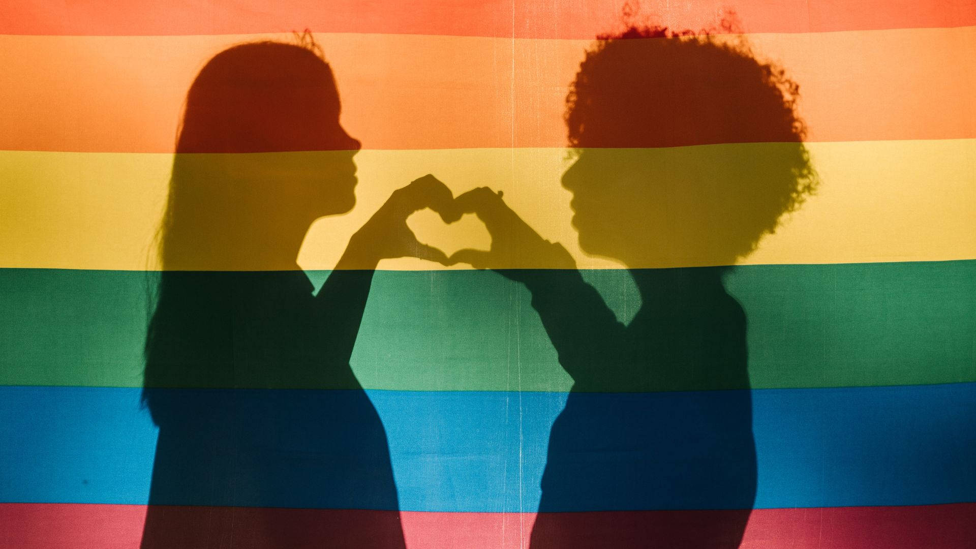 隱晦的LGBT女性剪影 壁紙 — 兩個剪影人物在鮮豔的彩虹旗背景下用手比出心形。