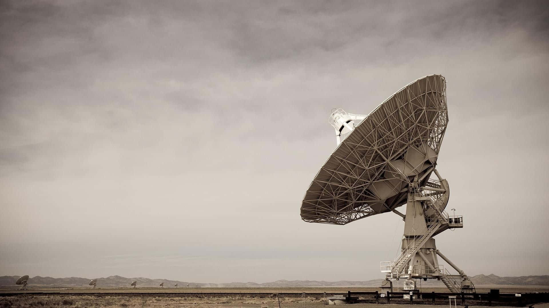 雄偉射電望遠鏡掃描星空 壁紙 — 一個大型衛星天線獨自矗立在廣闊的沙漠中，天空多雲，遠山可見。