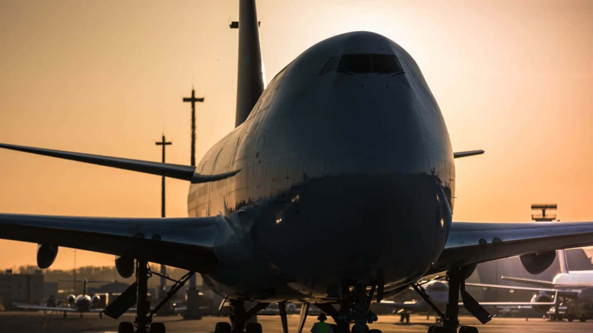 雄偉巨型客機高飛雲層之上 壁紙 — 一架巨大的飛機在溫暖的日落天空下剪影，位於機場停機坪上，遠處有機庫。