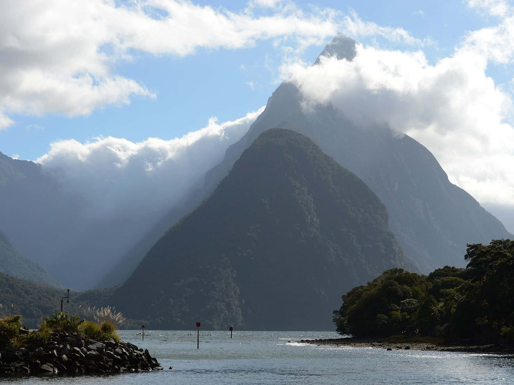 New Zealand 桌布 壁紙 — 雄偉的山峰穿過蓬鬆的白雲，俯瞰著被茂密綠色森林環繞的寧靜藍色海灣。