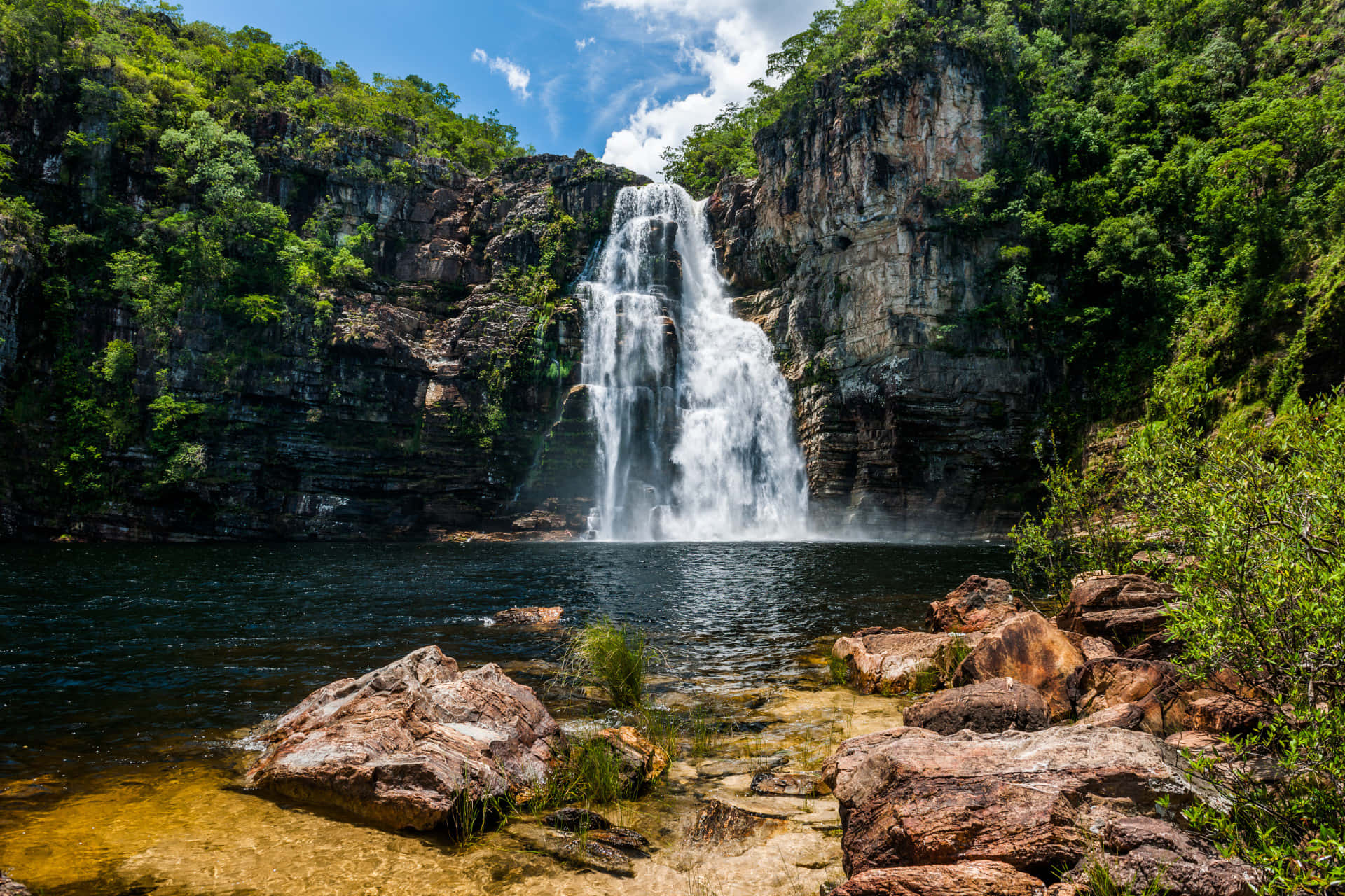 Eco-tourism 桌布 壁紙 — 雄偉的瀑布從崎嶇的懸崖傾瀉而下，流入一個被茂密綠植環繞的寧靜水潭，上方是明亮的藍天。