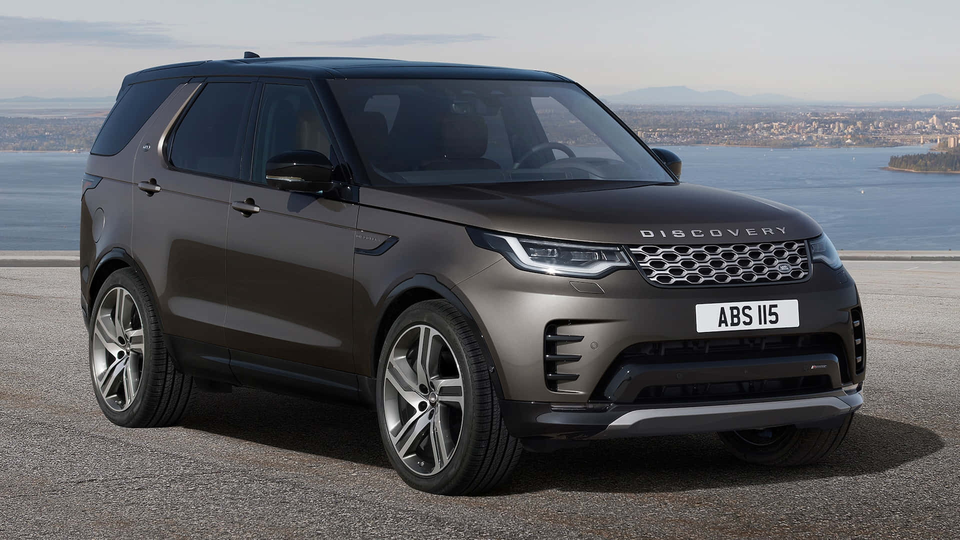 雄偉的路虎發現車探索荒野 壁紙 — 一架時尚的啞光棕色 Land Rover Discovery 停泊在海岸觀景點，背景是城市景觀和水面。