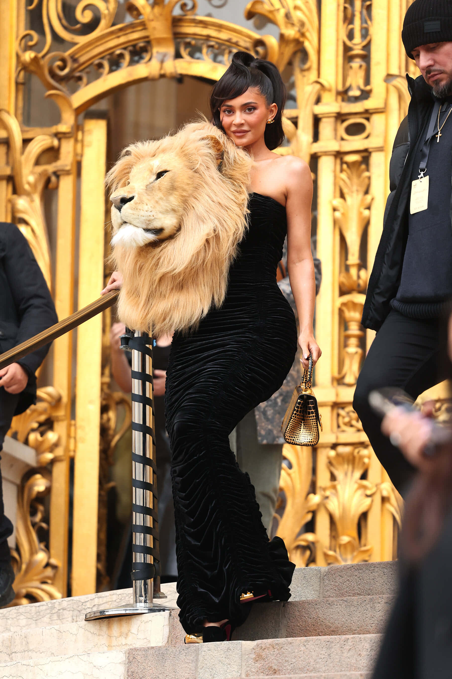雄獅頭像 壁紙 — Kylie Jenner 身穿黑色天鵝絨長裙，在華麗的金色大門旁，伴隨著獅頭道具走下樓梯。