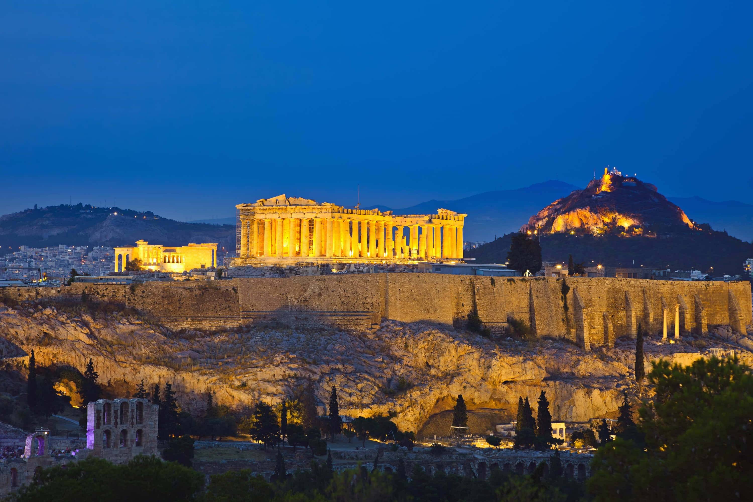 greek 桌布 壁紙 — 雅典衛城的帕德嫩神廟在暮色燈光下溫暖地發光，映襯著深藍色的天空和遠方的山丘。