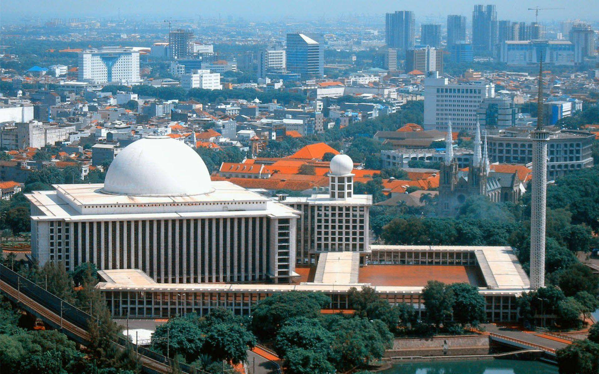 雅加達 Istiqlal 清真寺 壁紙 — 從高空俯瞰，一座宏偉的白色圓頂清真寺，帶有宣禮塔，矗立在密集的現代摩天大樓和紅頂建築組成的城市叢林中，籠罩在朦朧的日光下。