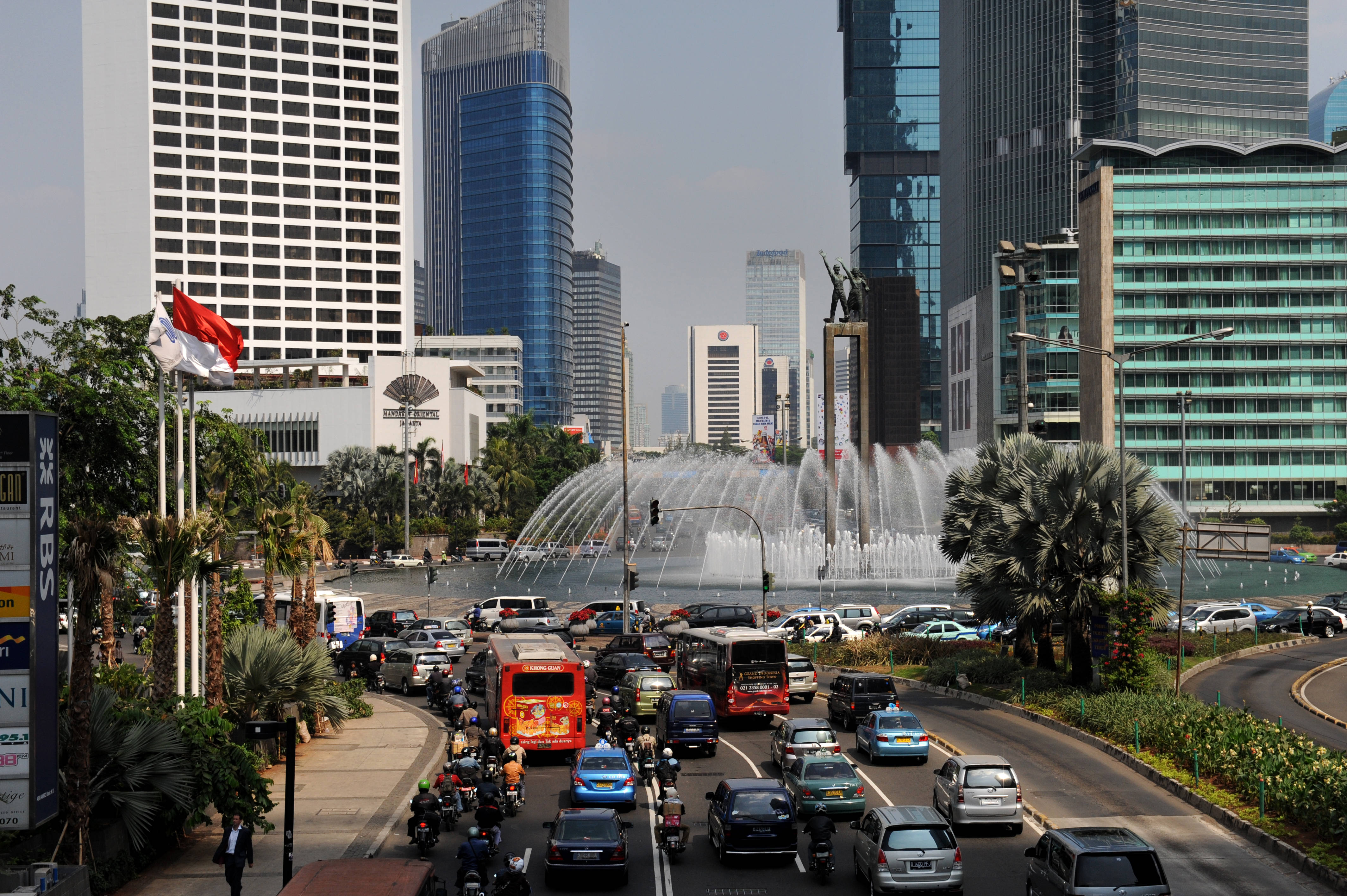 雅加達中心噴泉 壁紙 — 一條繁忙的城市街道，汽車和電單車川流不息，經過一個被現代摩天大樓和棕櫚樹環繞的大型噴泉。