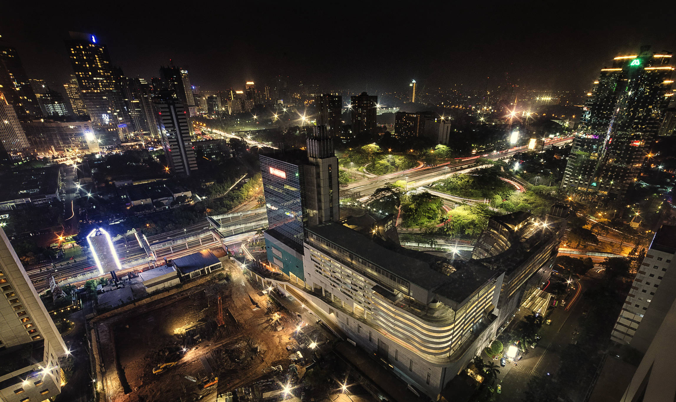 雅加達夜景燈光 壁紙 — 繁忙城市景觀的夜間鳥瞰圖，閃爍的摩天大樓和照明的高速公路蜿蜒穿過密集的城市建築。