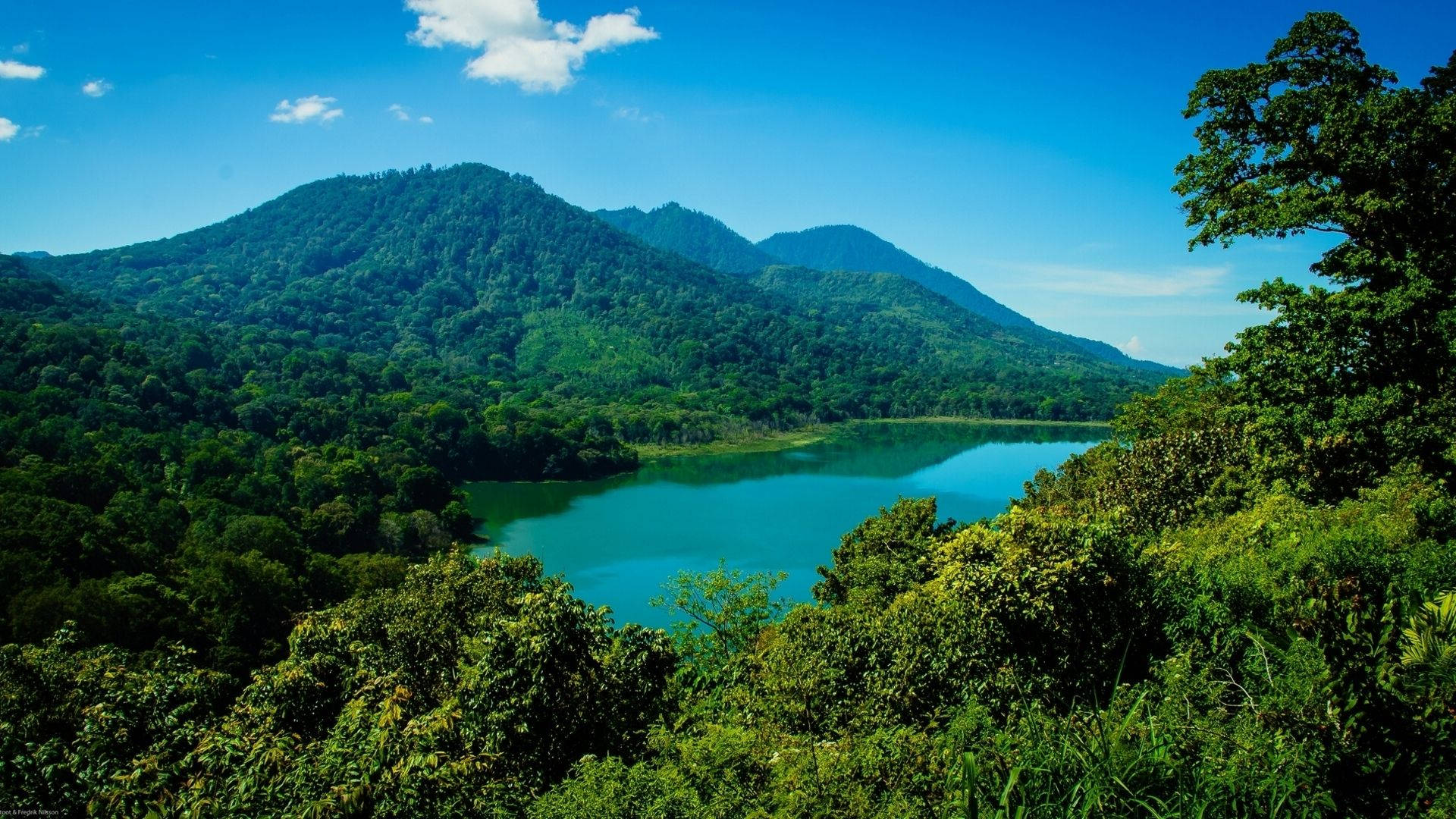 雅加達山景 壁紙 — 一個寧靜的藍綠色湖泊，坐落在鬱鬱蔥蔥的綠色山脈之間，上方是明亮的藍天，點綴著幾朵雲彩。