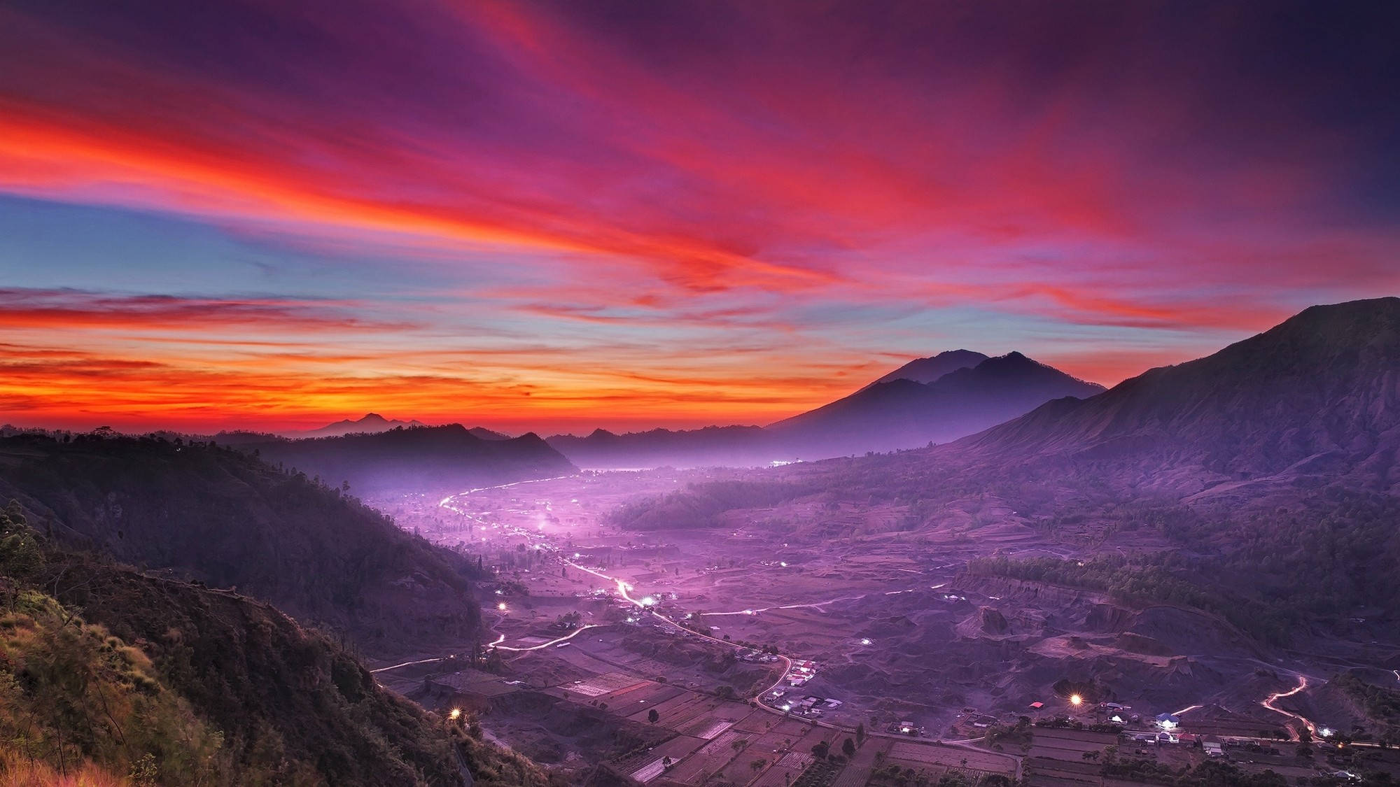 雅加達紫山 壁紙 — 充滿活力的日落將山脈和山谷染成火紅、紫和橙色，下方燈光閃爍。