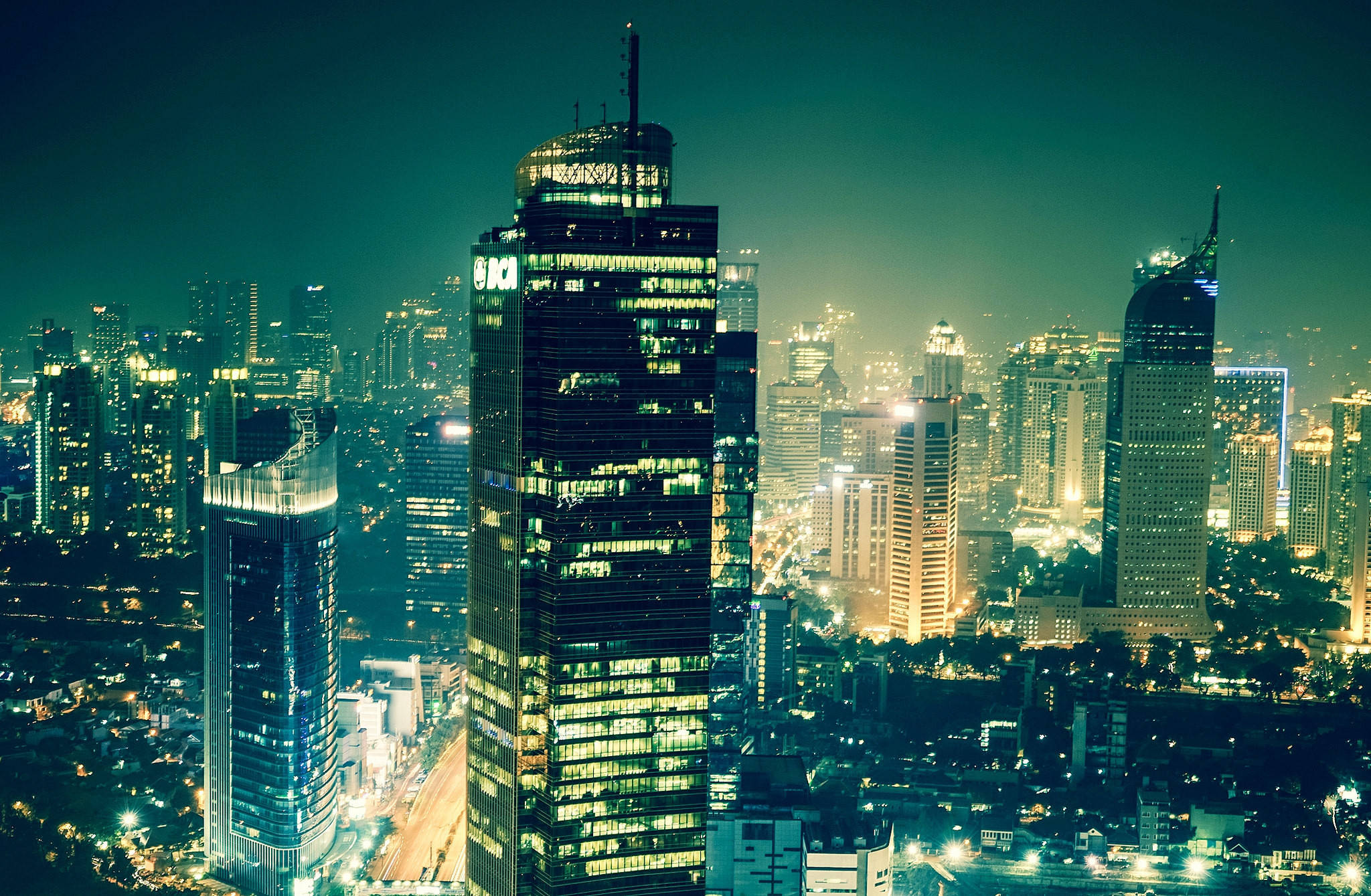 雅加達辦公大樓 壁紙 — 充滿活力的夜景城市景觀，高聳的摩天大樓在深藍綠色天空下閃耀。