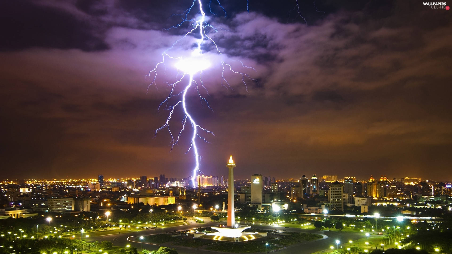 雅加達雷電交加 壁紙 — 一道巨大的閃電劃過夜間燈火通明的城市天際線，照亮了雲層和一座高大的紀念碑。