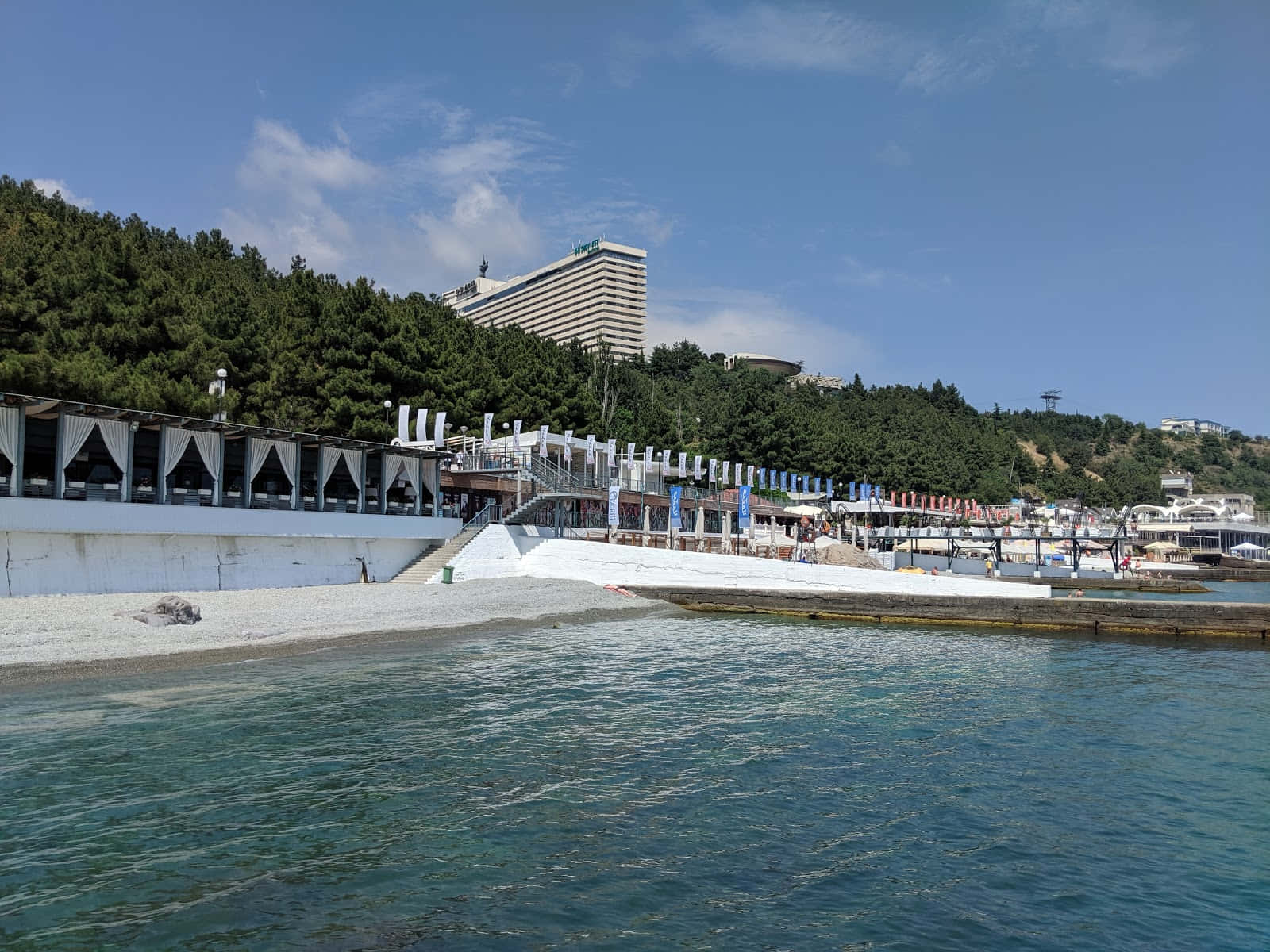 雅爾塔海灘景觀 壁紙 — 一個晴朗的日子，在海岸度假勝地，有碧綠的海水、溫柔的海浪、白色的卵石海灘和一座現代化酒店，坐落在綠色山丘之中。